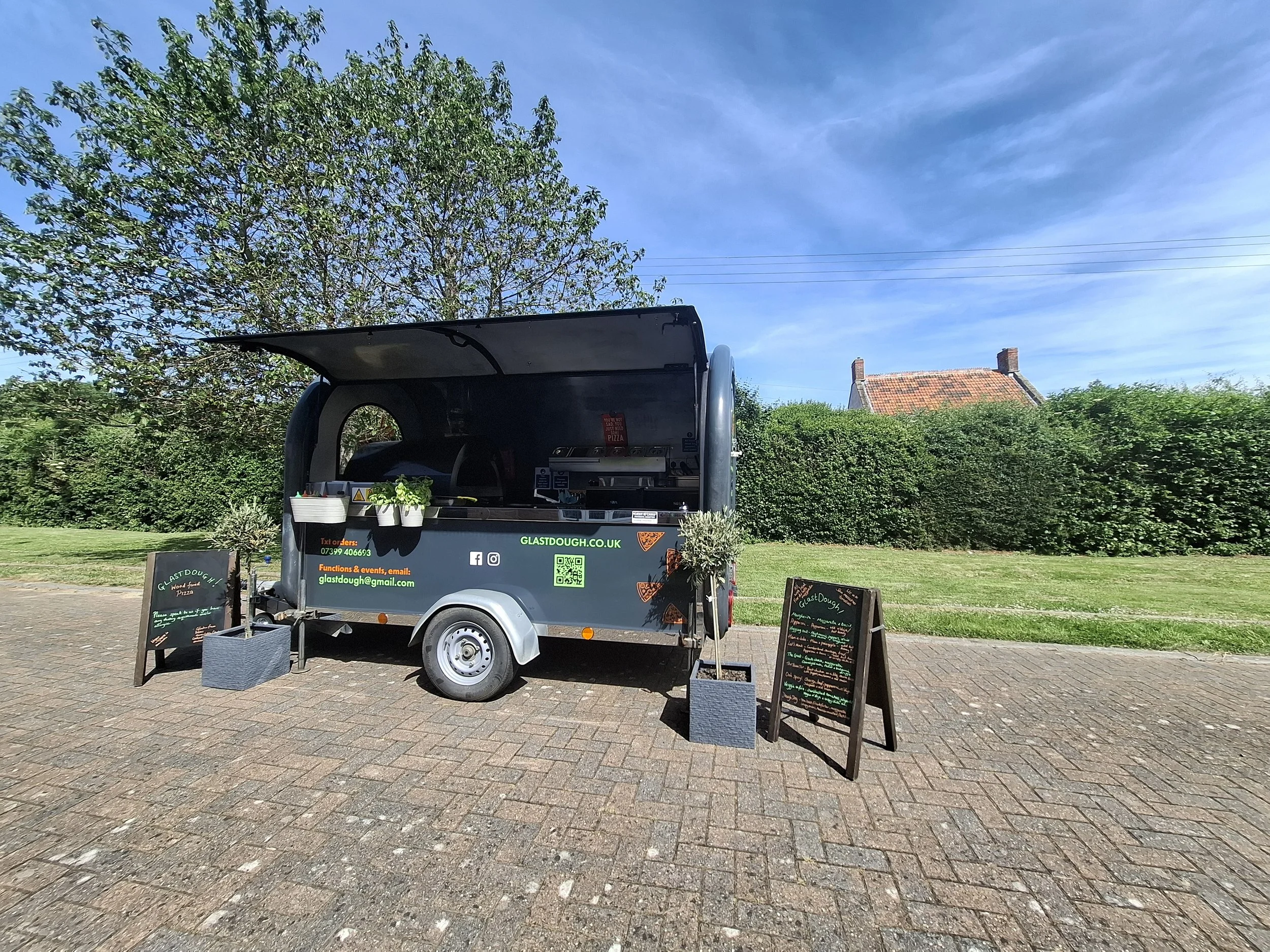A food truck with a black exterior and green text, parked on a brick-paved area with trees and a hedge in the background. Two chalkboard signs are displayed outside the truck, and the sky is blue with some clouds.