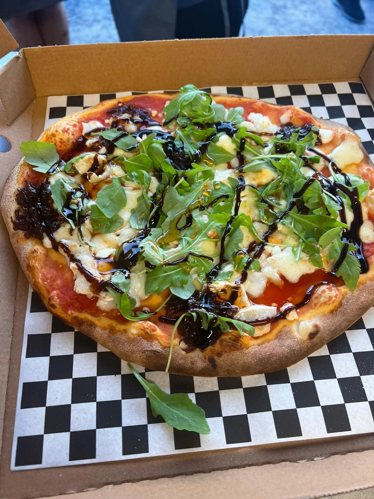 A pizza with cheese, arugula, and balsamic glaze in an open cardboard box on checkered paper.