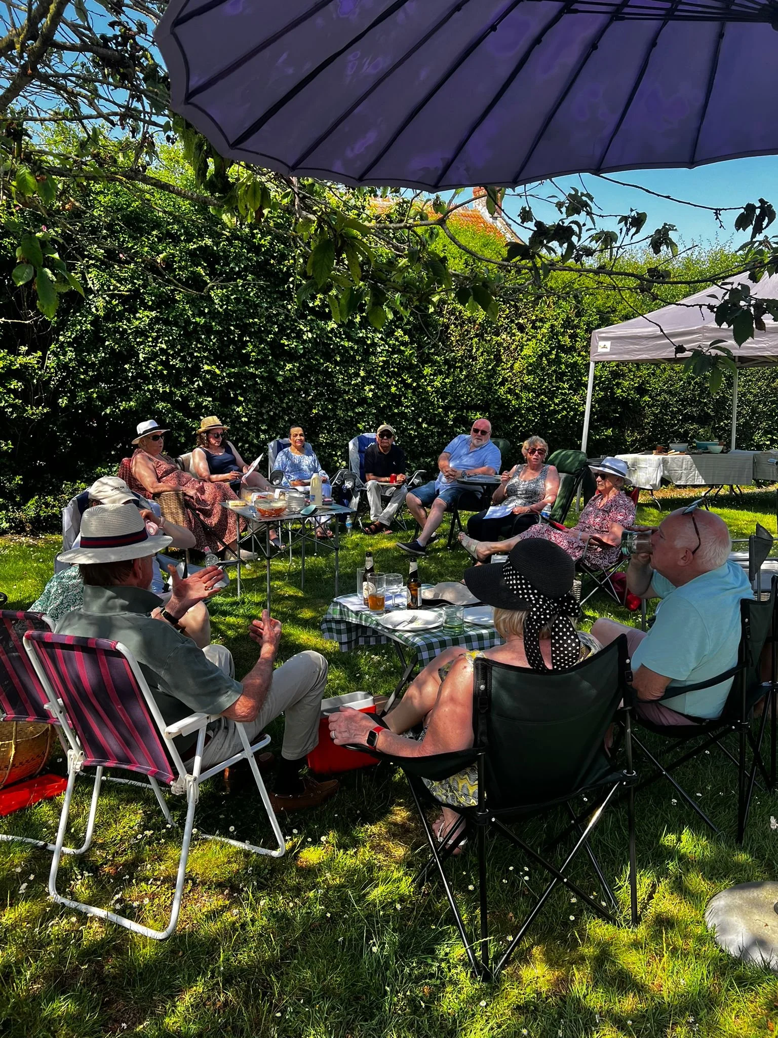 Group of people enjoying a sunny outdoor gathering in a garden, seated in chairs with drinks and snacks, under large umbrellas and trees.