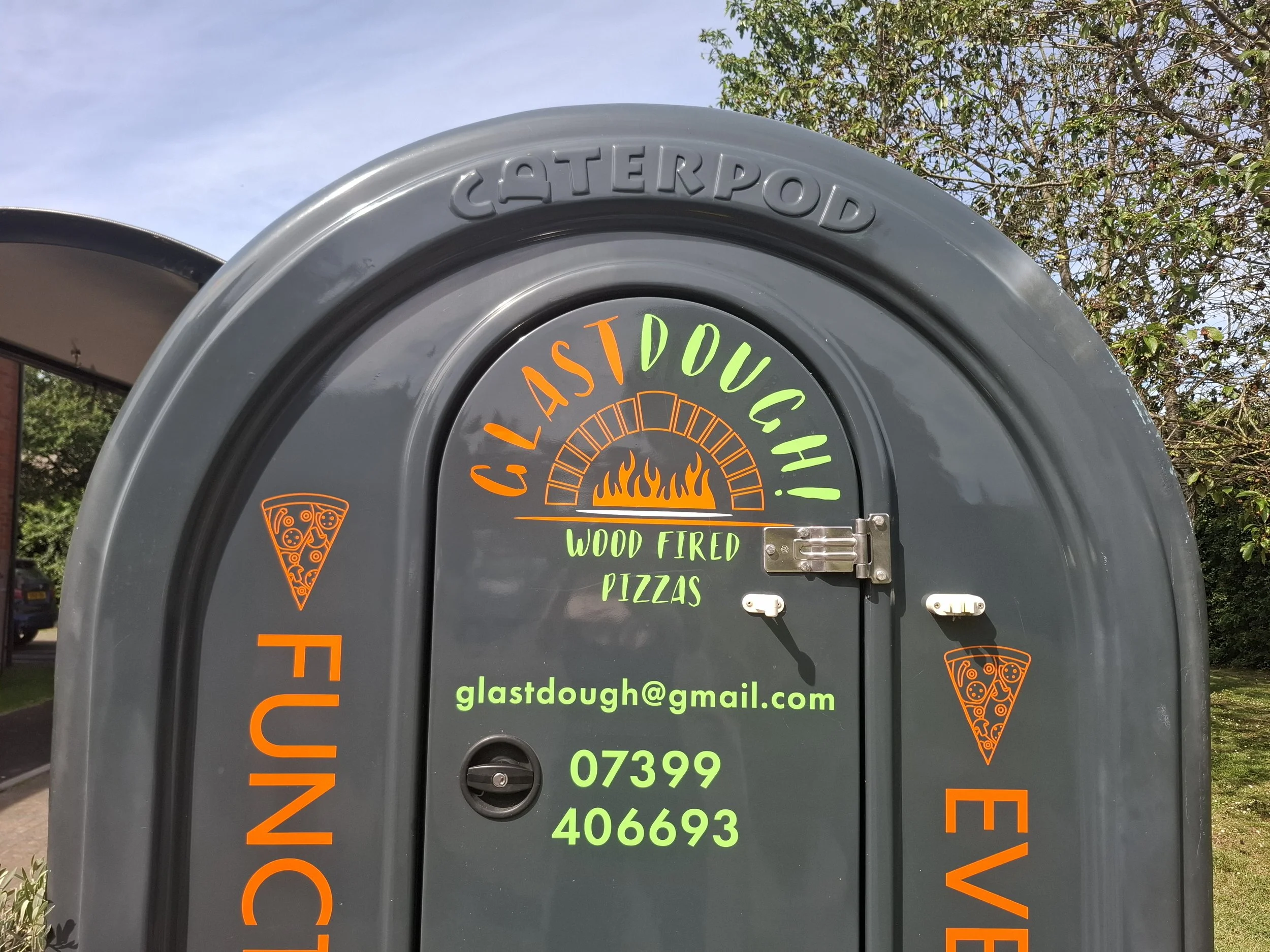 Grey outdoor sign advertising wood-fired pizzas from Glastdough, with orange and green text and pizza cone graphics, set against a background of trees and sky.