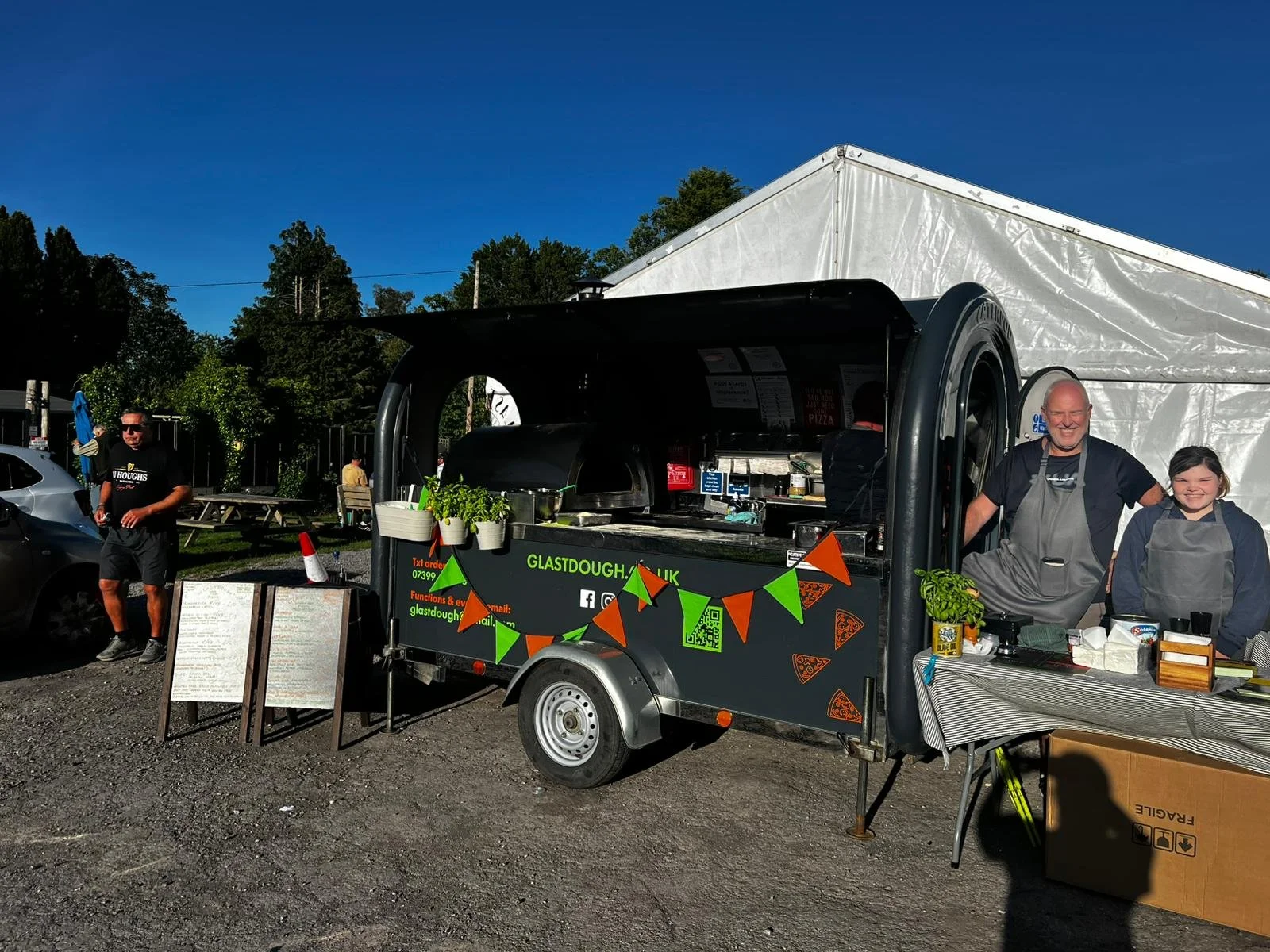A scene at an outdoor food stall, featuring a man and a young girl, both wearing aprons, standing behind a counter with various kitchen items and potted herbs. A man in sunglasses is walking nearby, with parked cars and a large white tent in the back