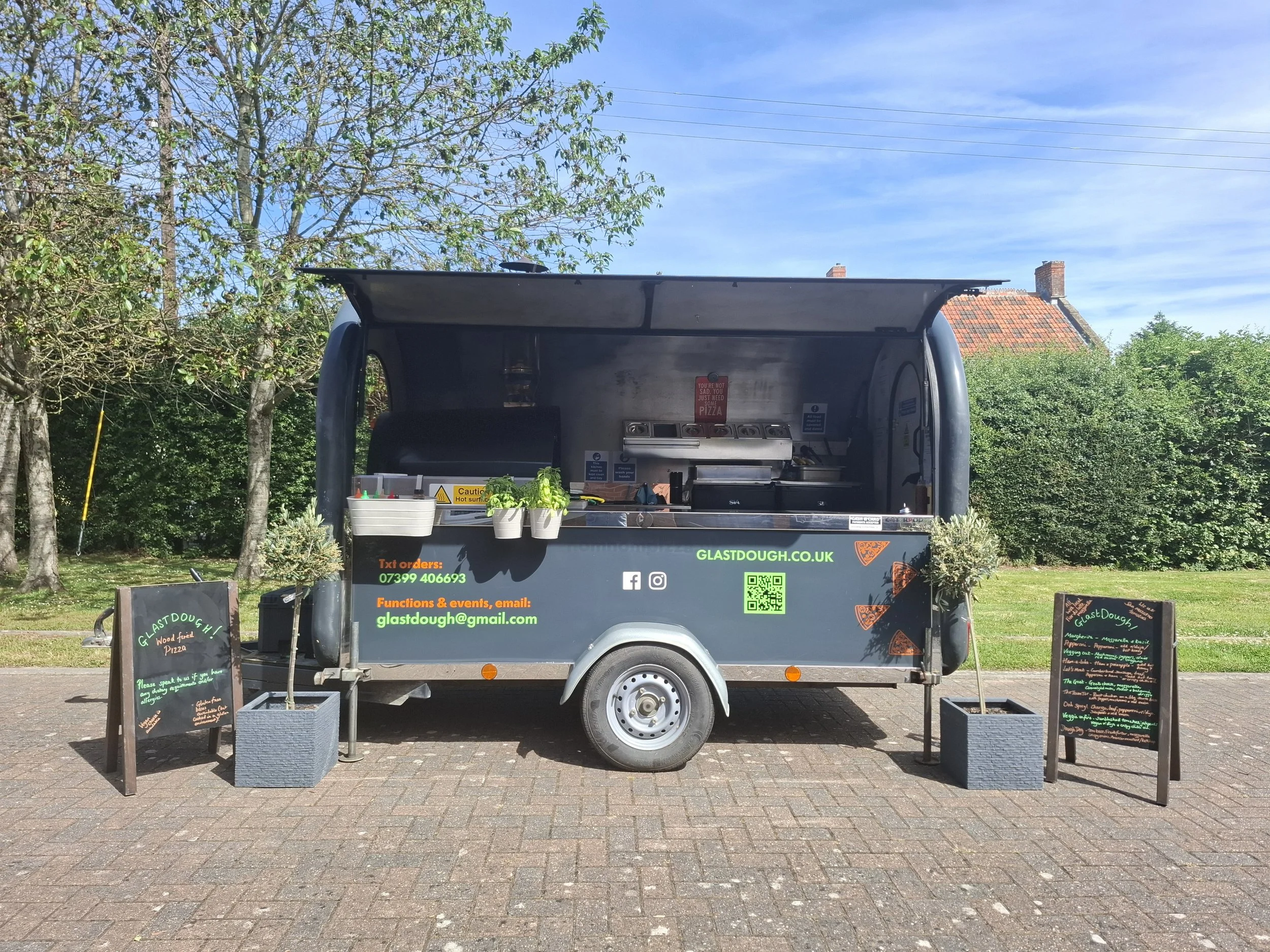 A food truck serving pizza, with menus displayed on chalkboards on either side. The truck is parked outdoors on a paved area, with trees and a house with a red roof in the background. The truck has various stickers, contact information, and a QR code.