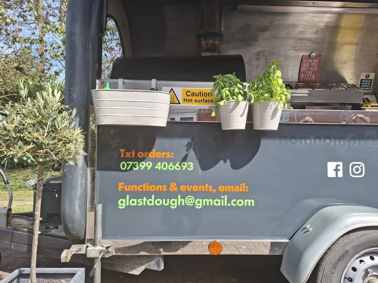 A food truck with a black exterior, featuring green and orange text displaying contact information and social media icons. Plants are attached to the side of the truck, and a caution sign warns of a hot surface.