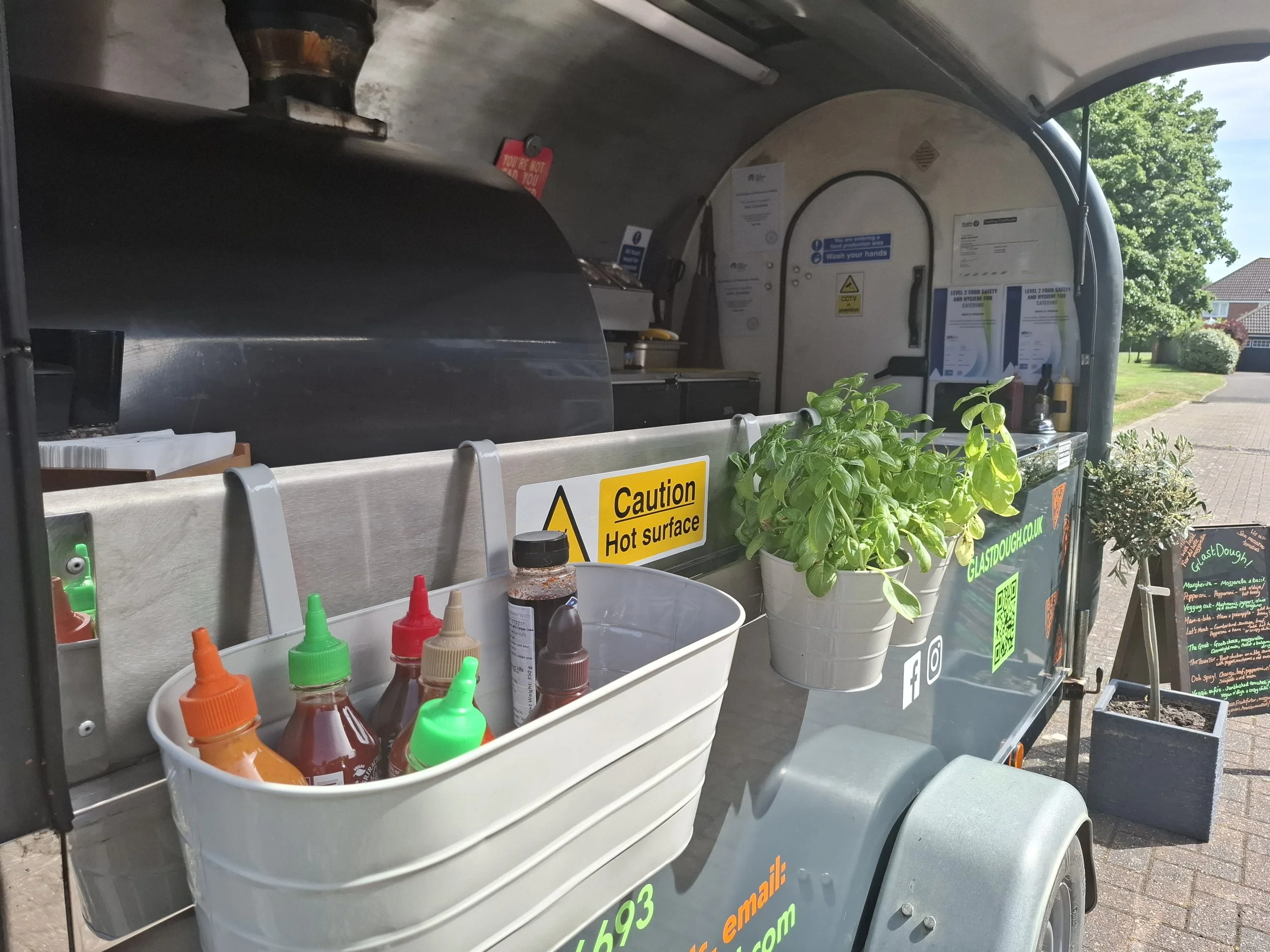 A food cart selling hot sauce with a bunch of basil plants in pots, various labeled sauces, and signs warning about hot surface and promoting handwashing, located outdoors on a sunny day.