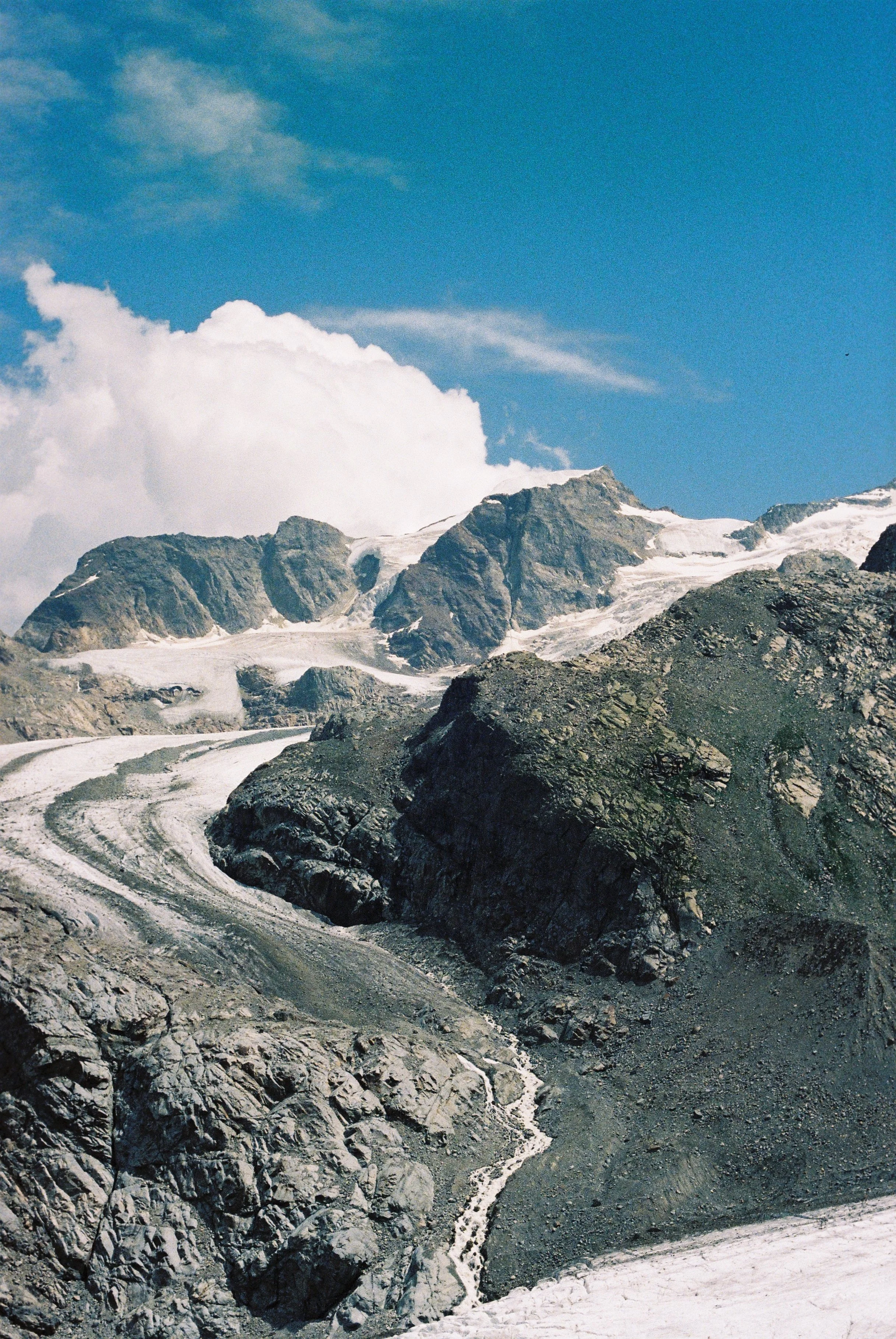 Snow-covered mountain peaks with rocky slopes and a winding snow-covered path in the foreground under a blue sky with white clouds.
