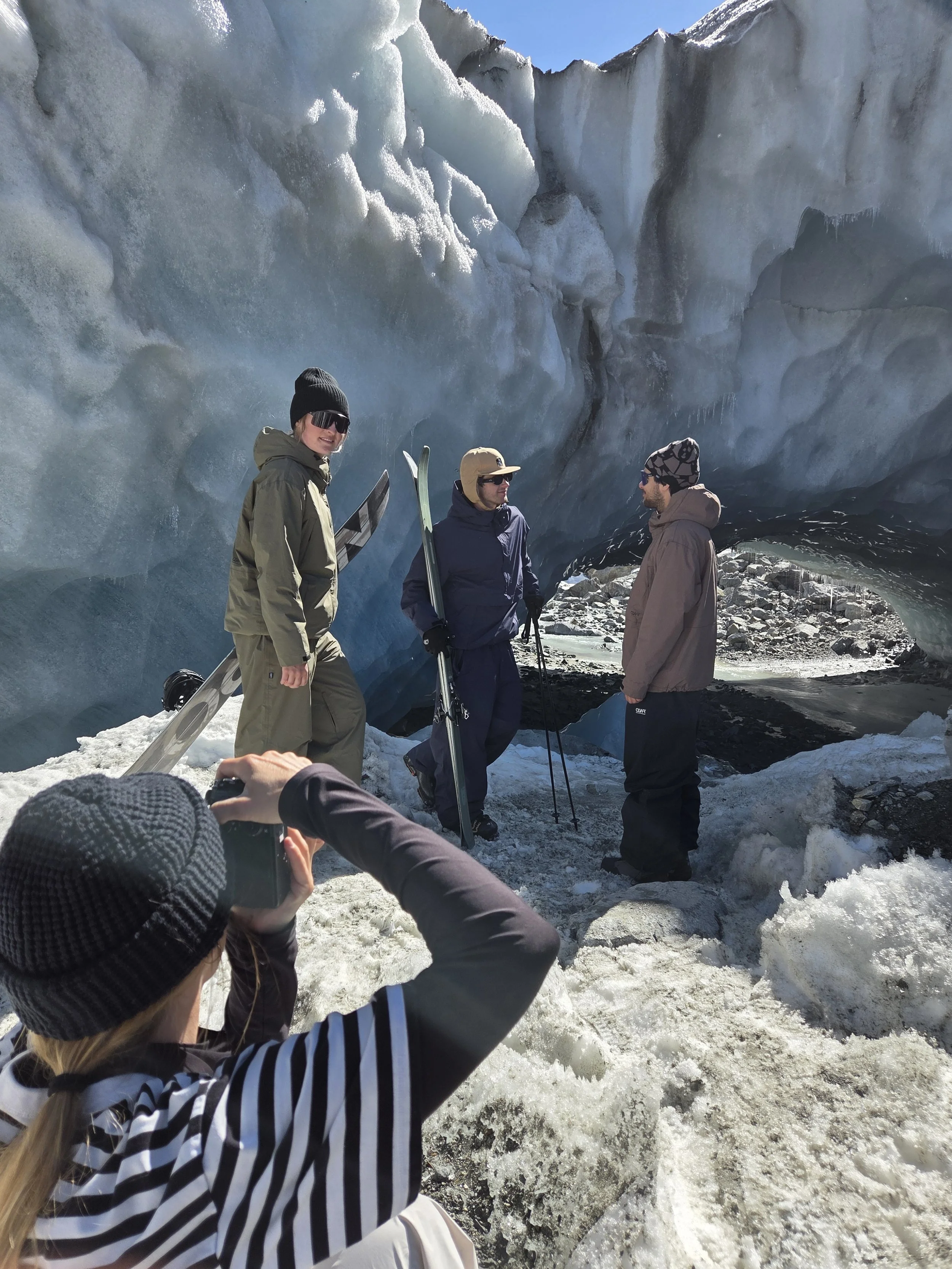Three people standing in front of a large ice formation, with one person taking a photo of the scene. The ice is blue and white, with some icicles hanging from the top.