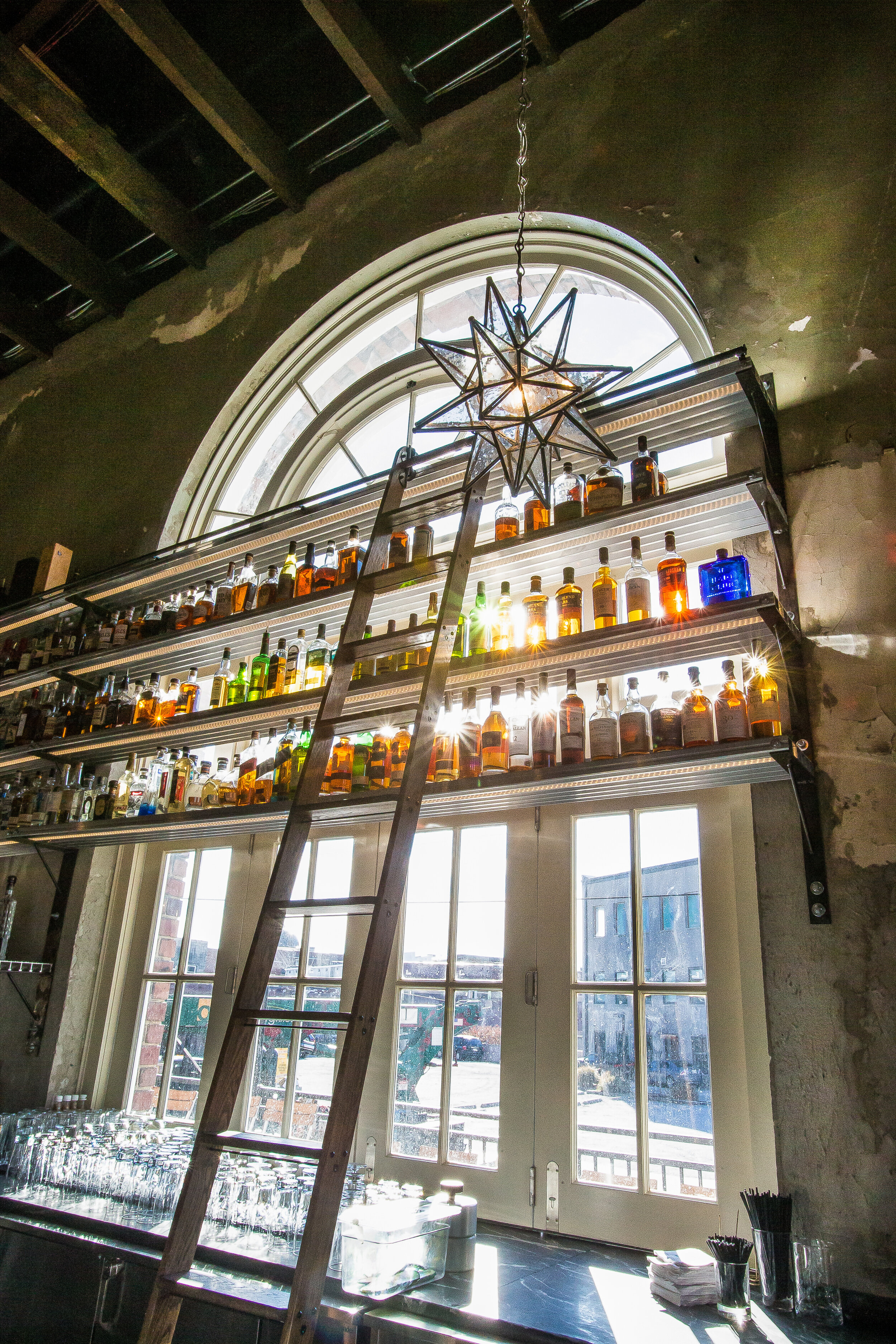 Interior of a bar or restaurant with a large arched window, a decorative star-shaped hanging light, and shelves holding bottles of various spirits. A ladder leans against the shelves, with sunlight streaming through the window.