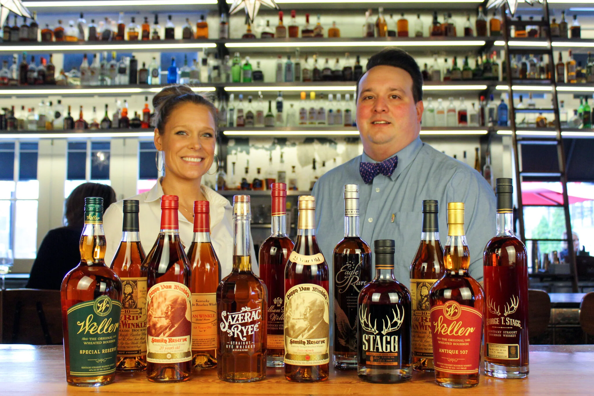 A bar with a display of whiskey bottles in front of two smiling bartenders, a woman and a man, with shelves of liquor bottles behind them.