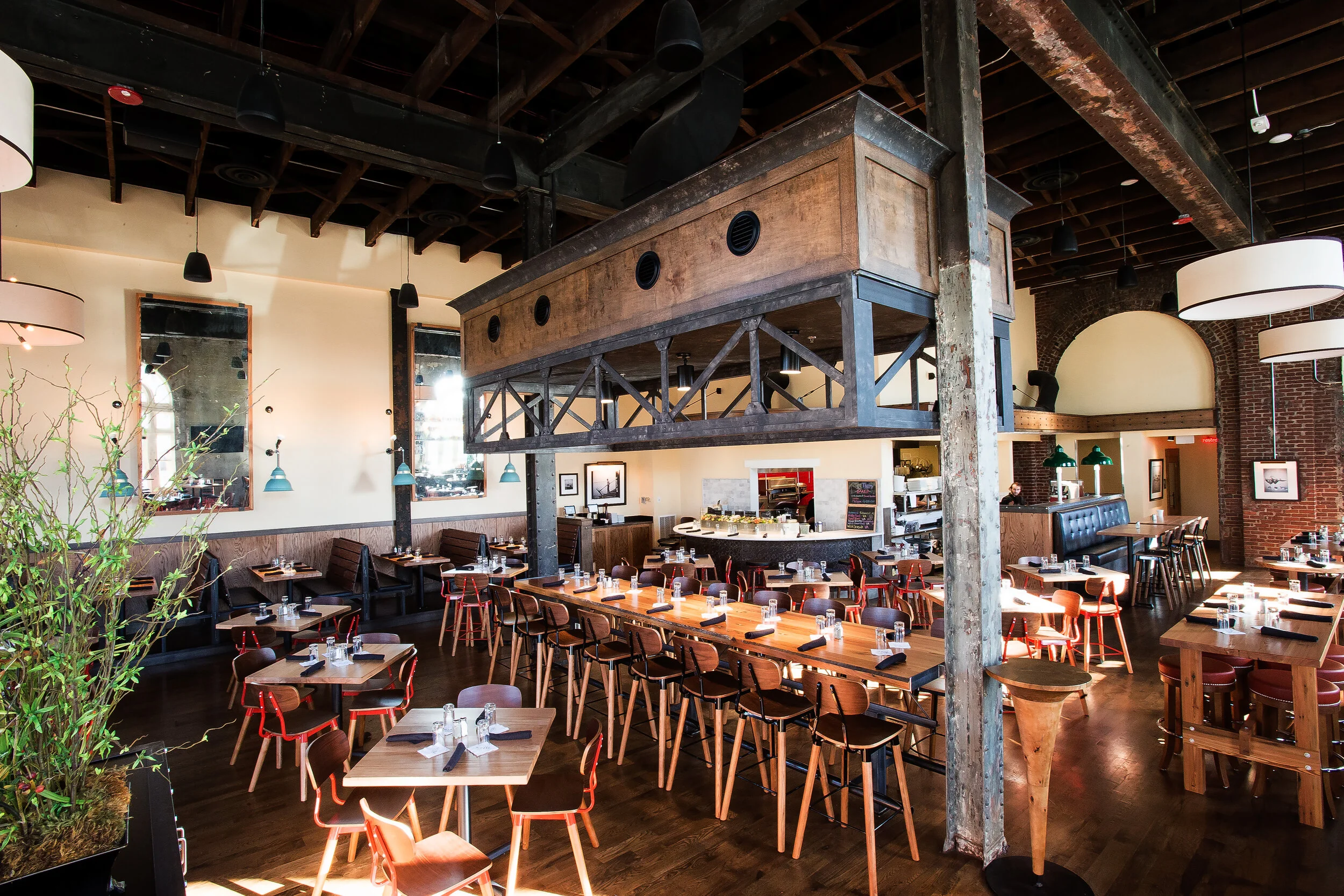 Interior of a restaurant with wooden tables and chairs, large hanging lights, mirrors on the walls, and exposed brick and wooden ceiling beams.