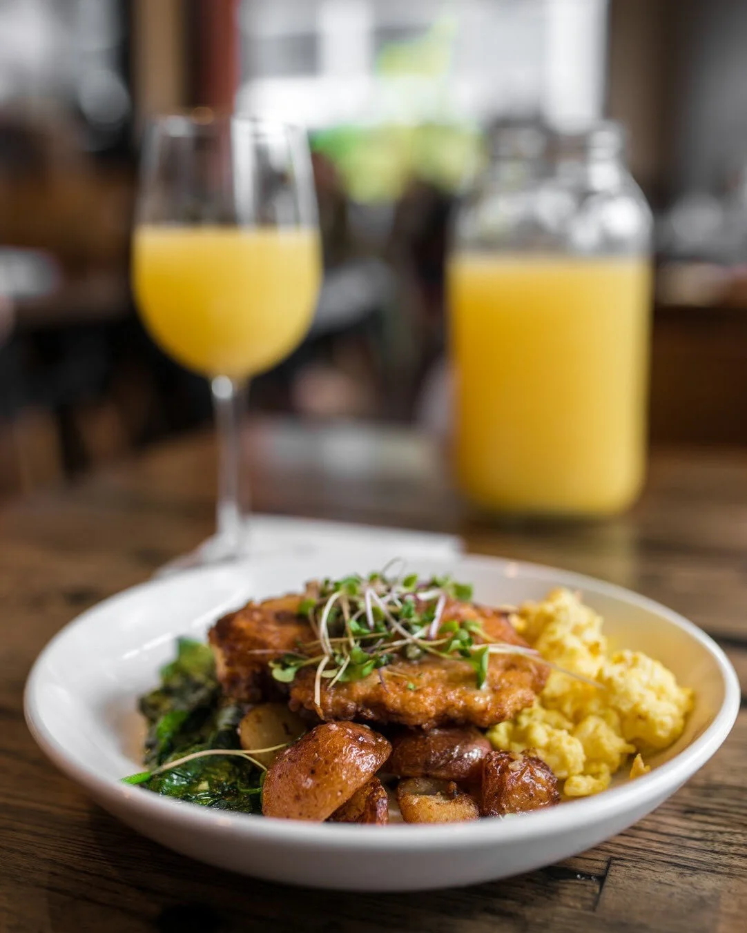 A white plate with a serving of scrambled eggs, roasted potatoes, a cooked piece of chicken with sprouts on top, and greens, placed on a wooden table. In the background, a glass of orange juice and a jar of more orange juice are visible, with a blurr