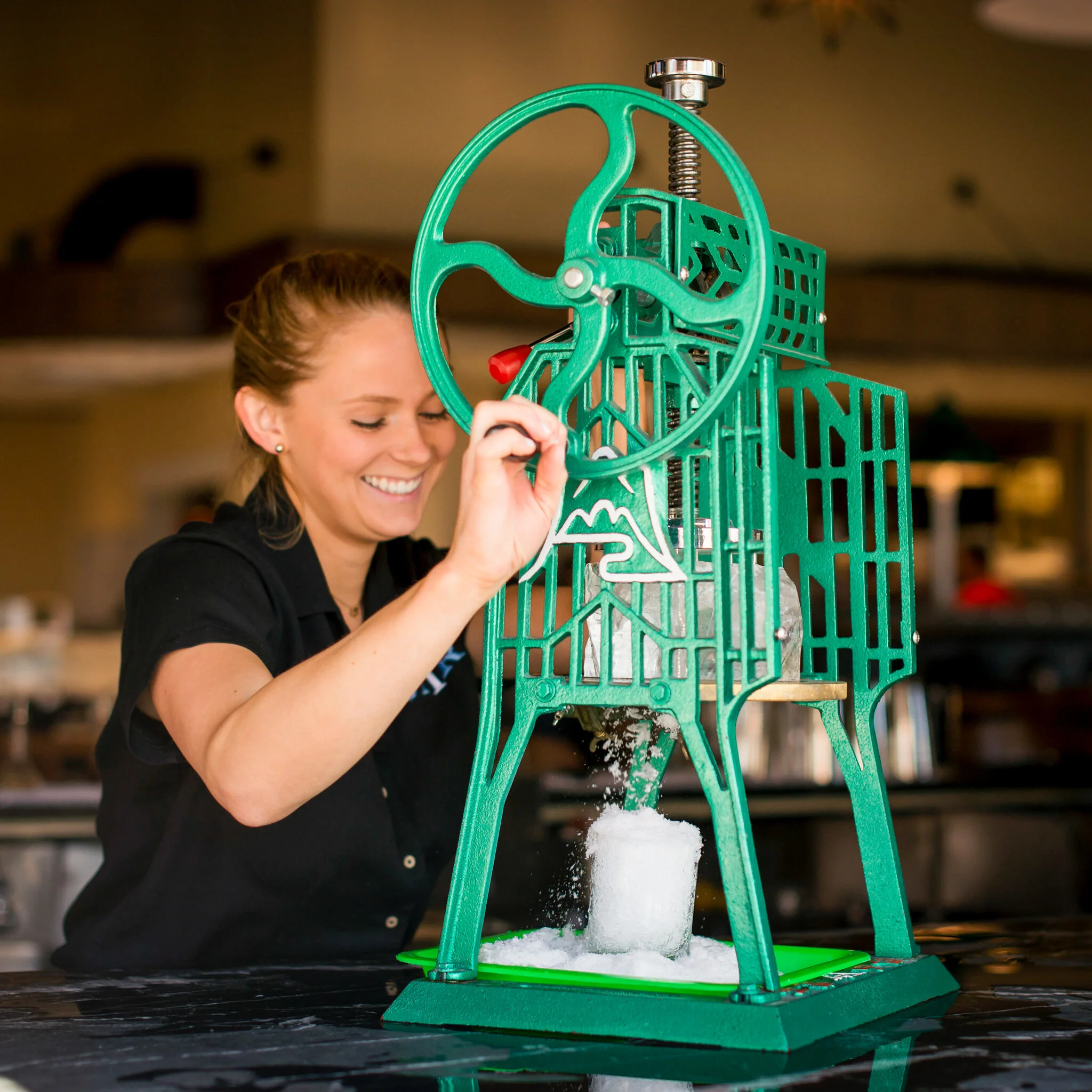 Close-up of a person operating a green ice shaver machine, making snow cones or shaved ice with white snow falling from the machine.