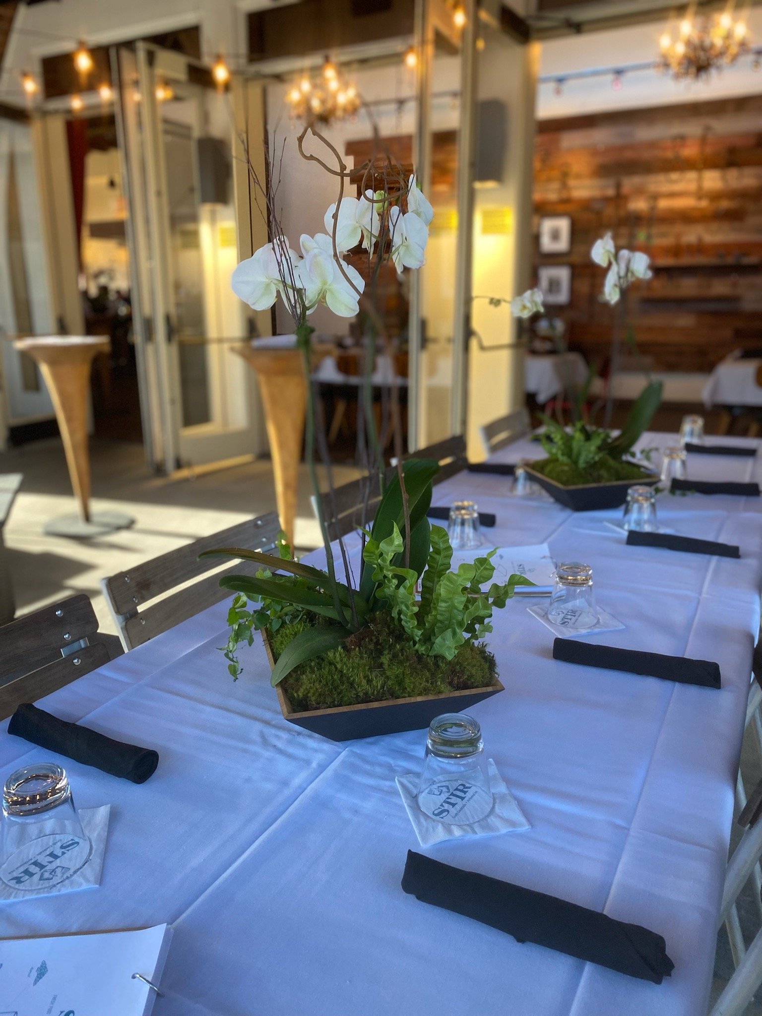 A dining table set for a meal features a floral arrangement with white orchids and greenery in a rectangular black container at the center. The table has a white tablecloth, black napkins, and clear glasses. The background shows a cozy restaurant wit