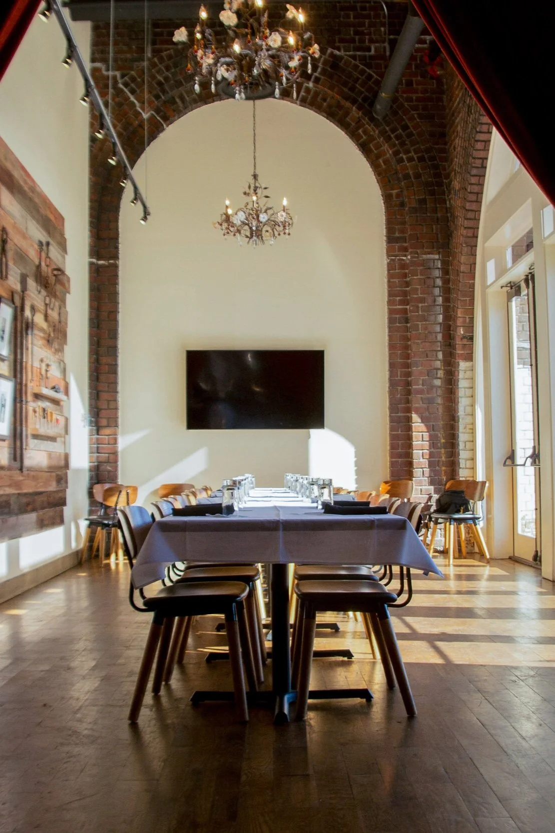 A long rectangular table set for a meeting or event inside a room with exposed brick walls and high ceilings. The table has white tablecloths, glasses, and placemats. The room features chandeliers, a large TV screen on the wall, and large windows let