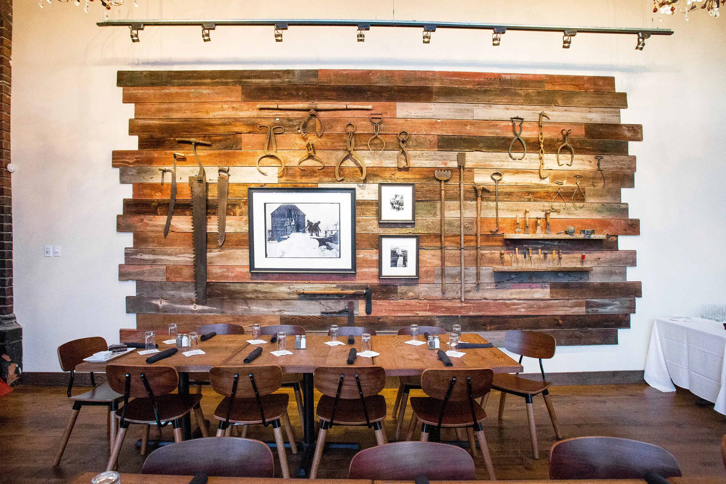 A rustic dining room features a wooden wall decorated with vintage farming tools and black and white photographs, with a large wooden table set for a meal.