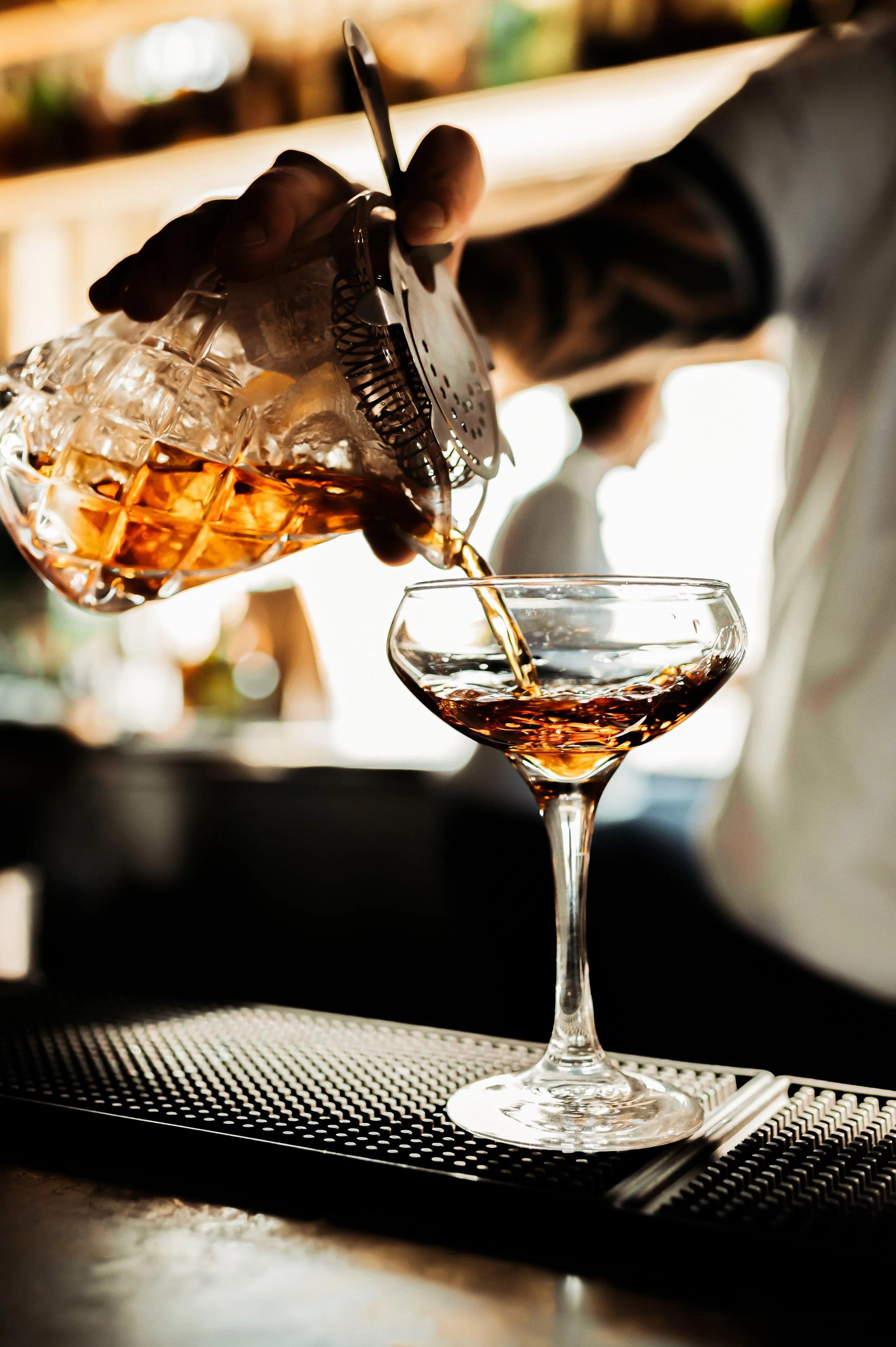 A bartender pours whiskey into a cocktail glass at a bar, with a blurred background and warm lighting.
