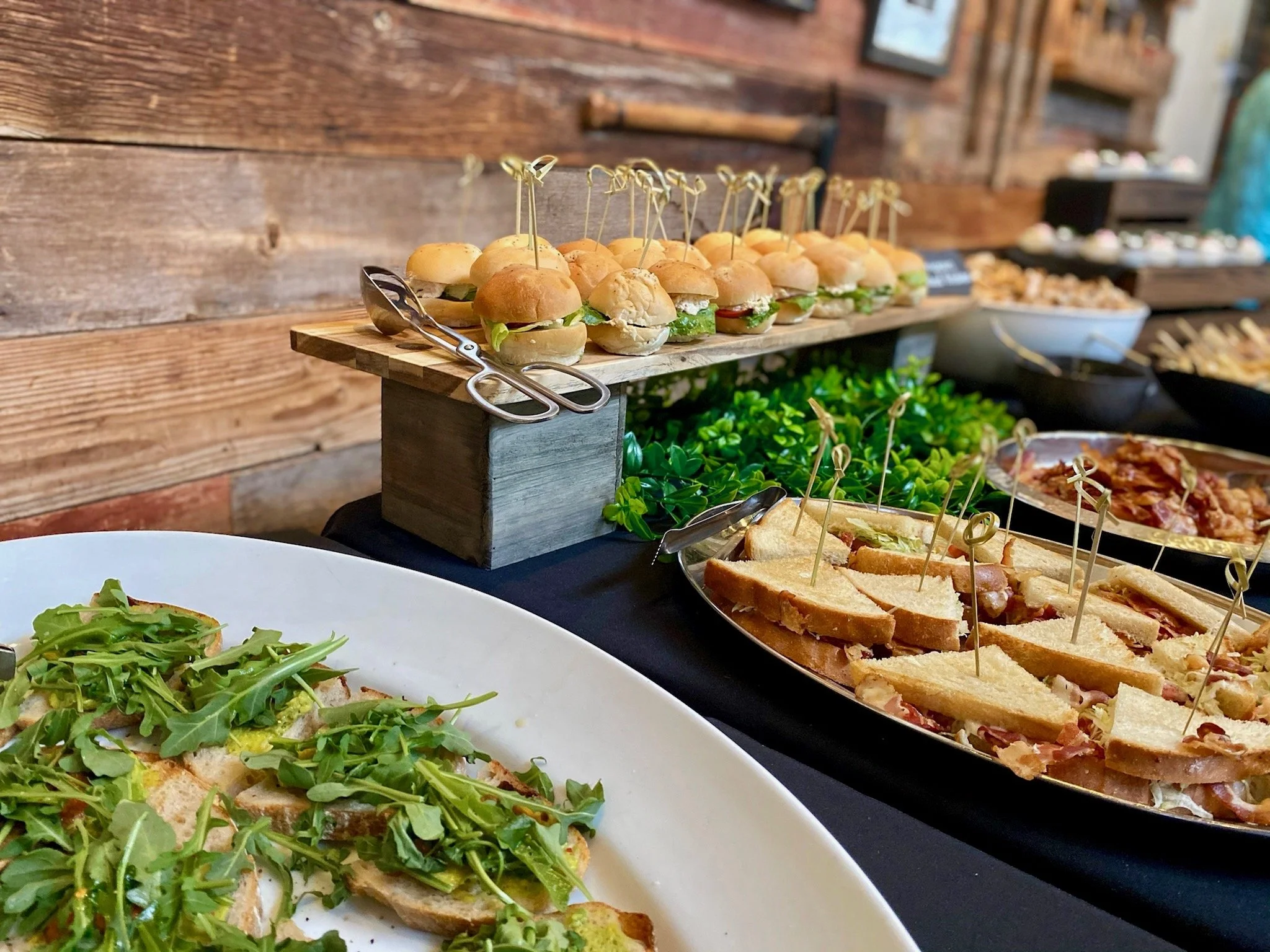 Tray of mini sandwiches with various fillings, skewered with decorative picks, on a wooden serving board. In the background, additional food items and a rustic wooden wall.