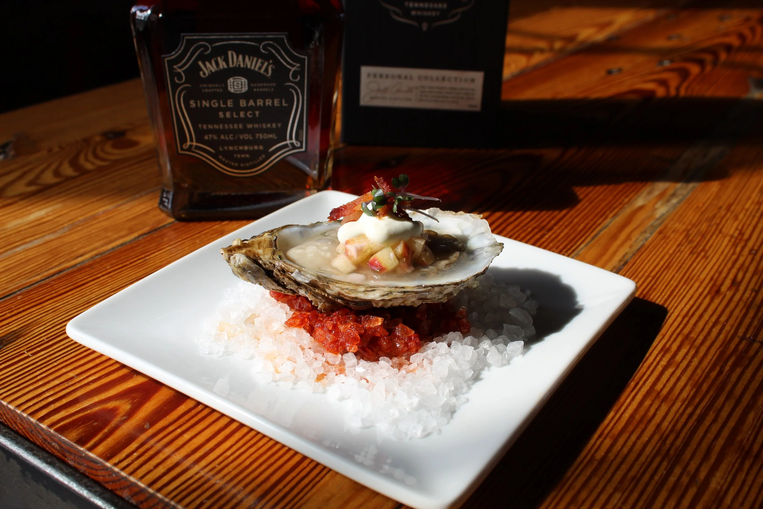 A gourmet oyster on a bed of ice served on a white square plate with a side of red sauce and garnished with microgreens, with a bottle of Jack Daniel's whiskey in the background on a wooden table.