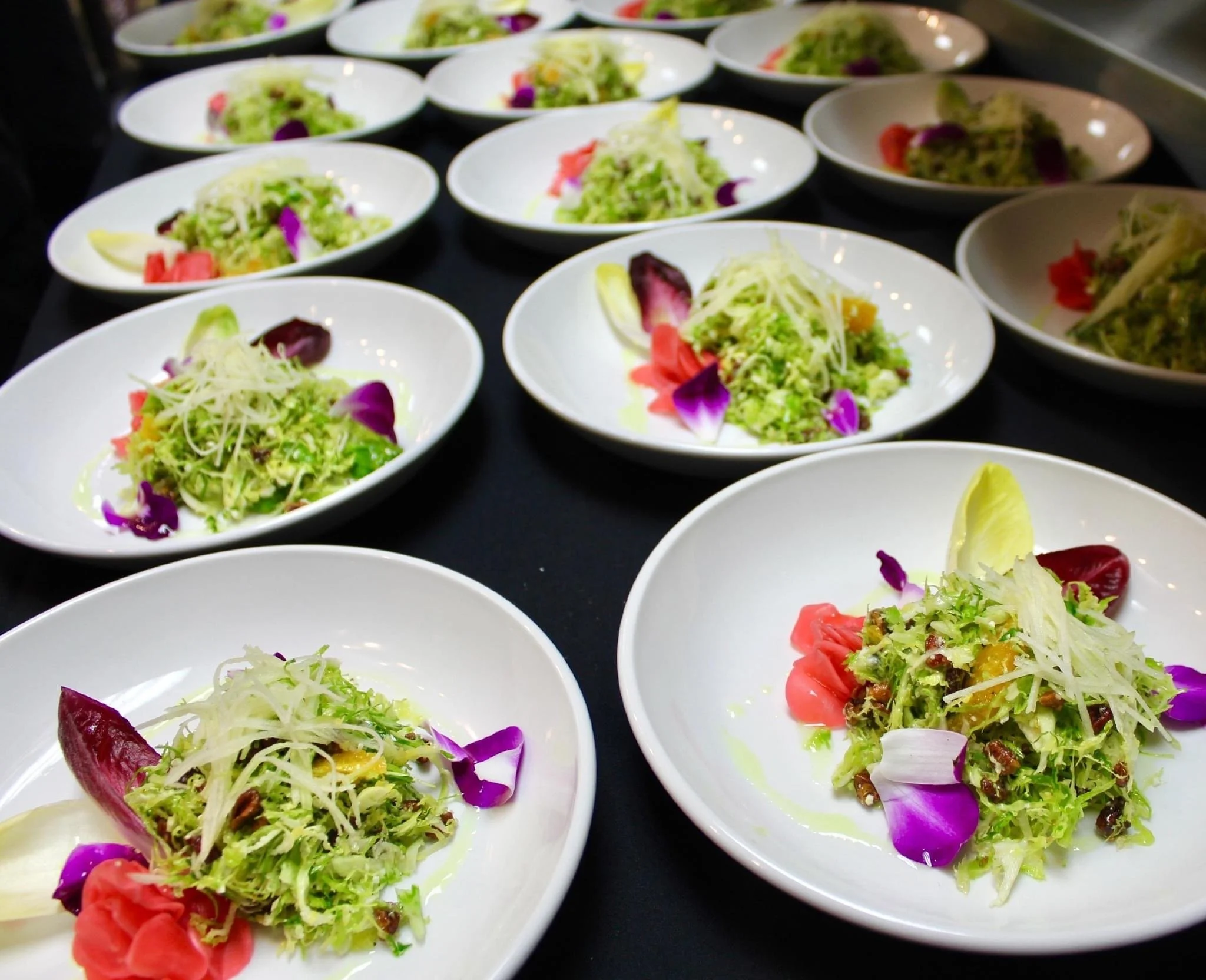Several white bowls filled with fresh mixed vegetable salads topped with edible flower petals on a dark surface.