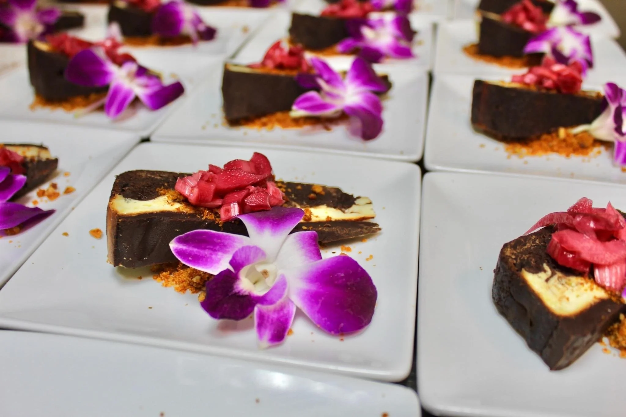 Square white plates with slices of chocolate cheesecake topped with pink flower petals and pink diced fruit, garnished with purple orchid flowers.