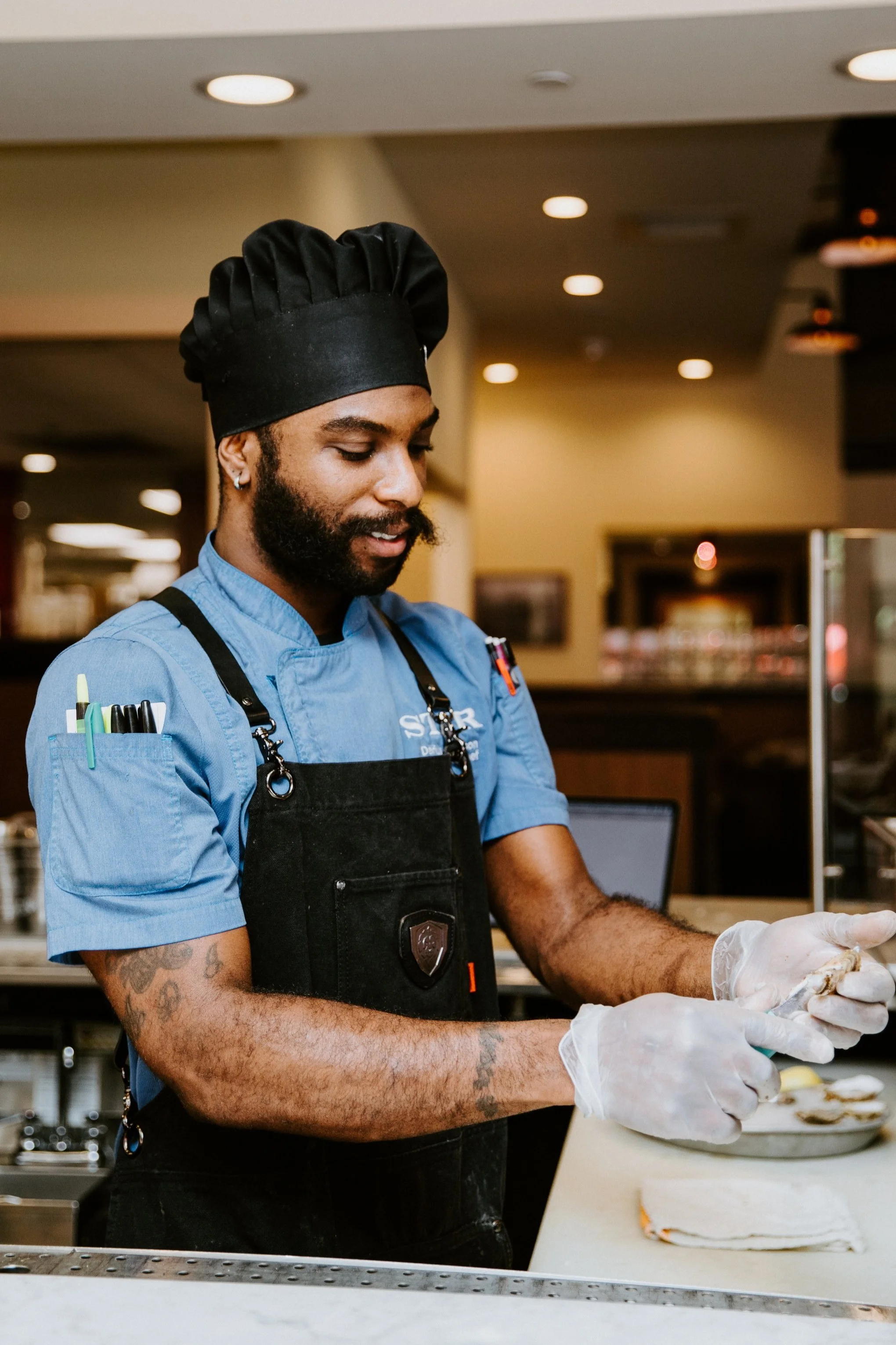A male chef with a black chef hat and earrings, wearing a blue uniform and black apron, preparing food in a restaurant kitchen. He has tattoos on his arms and is handling raw chicken with gloves.
