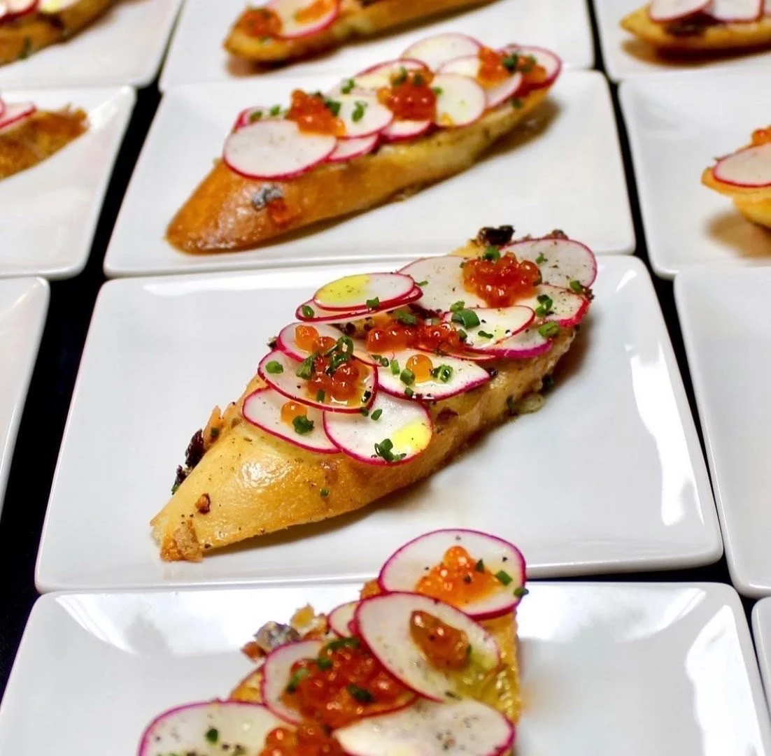 Several slices of open-faced sandwiches on white plates topped with radish slices, orange fish roe, chopped chives, and a drizzle of oil.