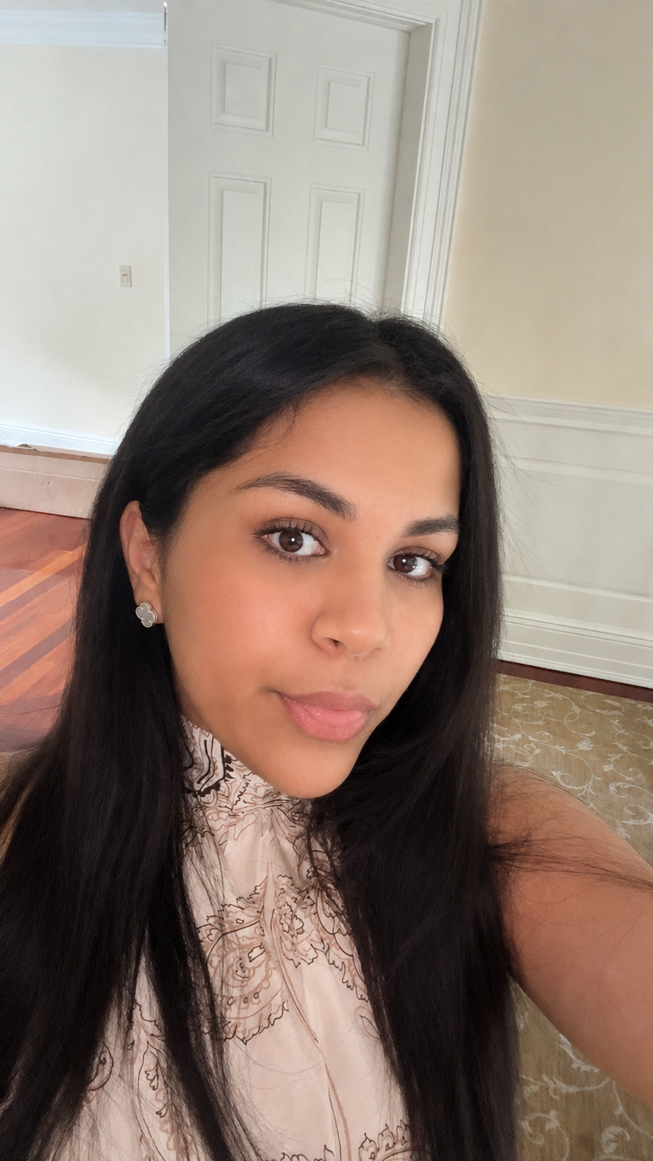 A young woman with long black hair and brown eyes taking a selfie indoors, wearing a cream-colored top with floral embroidery and silver earrings, with a white door, cream walls, hardwood flooring, and a patterned rug in the background.