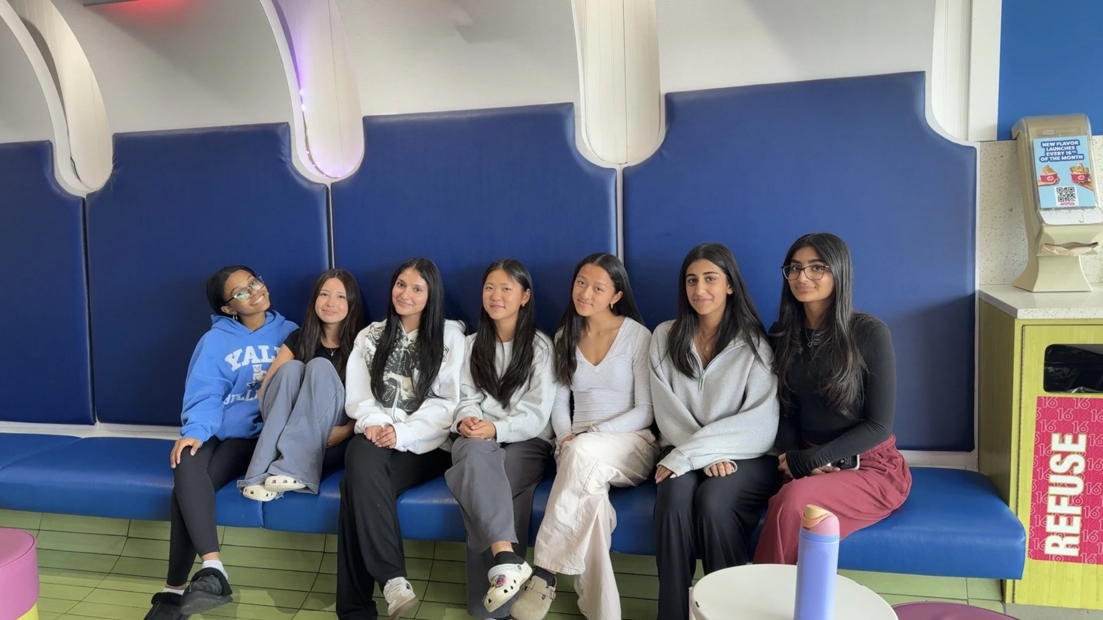 Six young women sitting on a blue bench inside an ice cream shop, smiling at the camera. The background features white walls with blue padded panels and a yellow ice cream dispenser to the right.