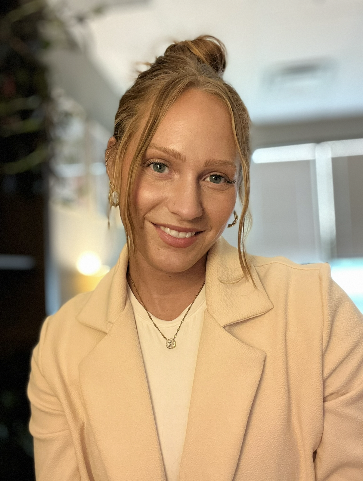 Close-up of a smiling woman with reddish hair and blue eyes, wearing a cream-colored blazer, a white top, gold jewelry, and standing in a well-lit indoor setting with a blurred background.