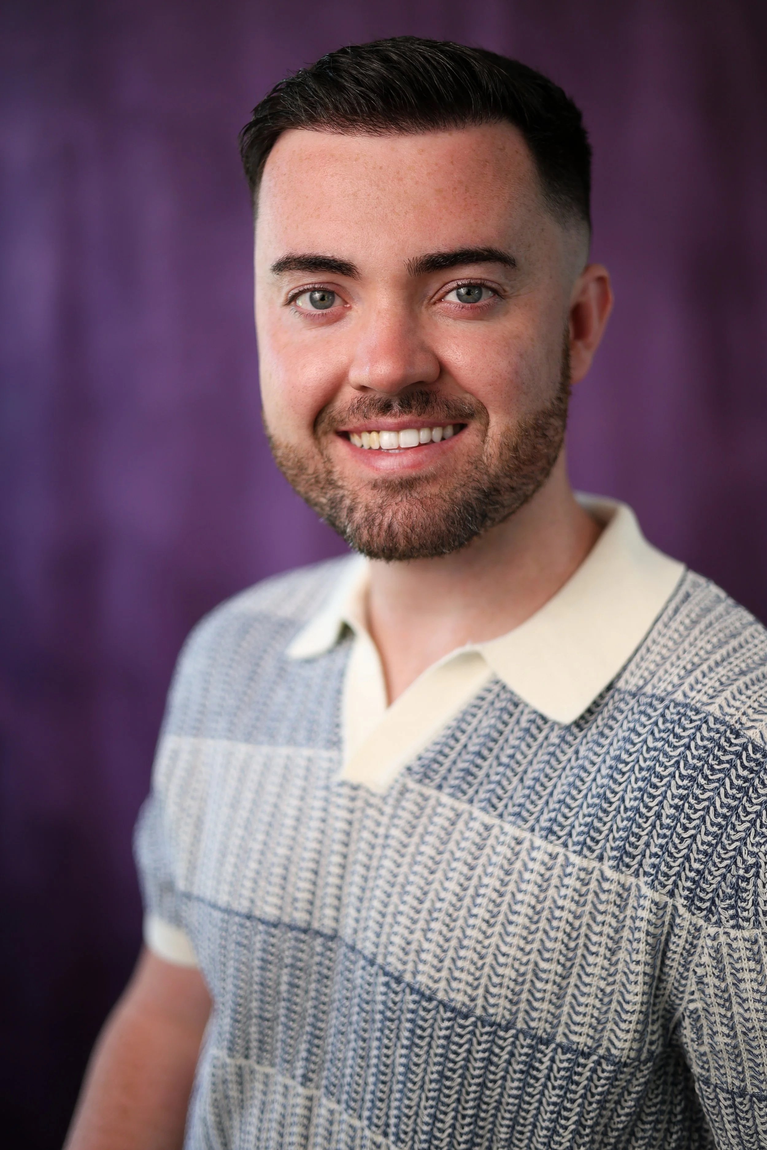 Close-up portrait of a smiling man with short dark hair, light skin, and a beard, wearing a patterned sweater over a white collared shirt, against a purple background.