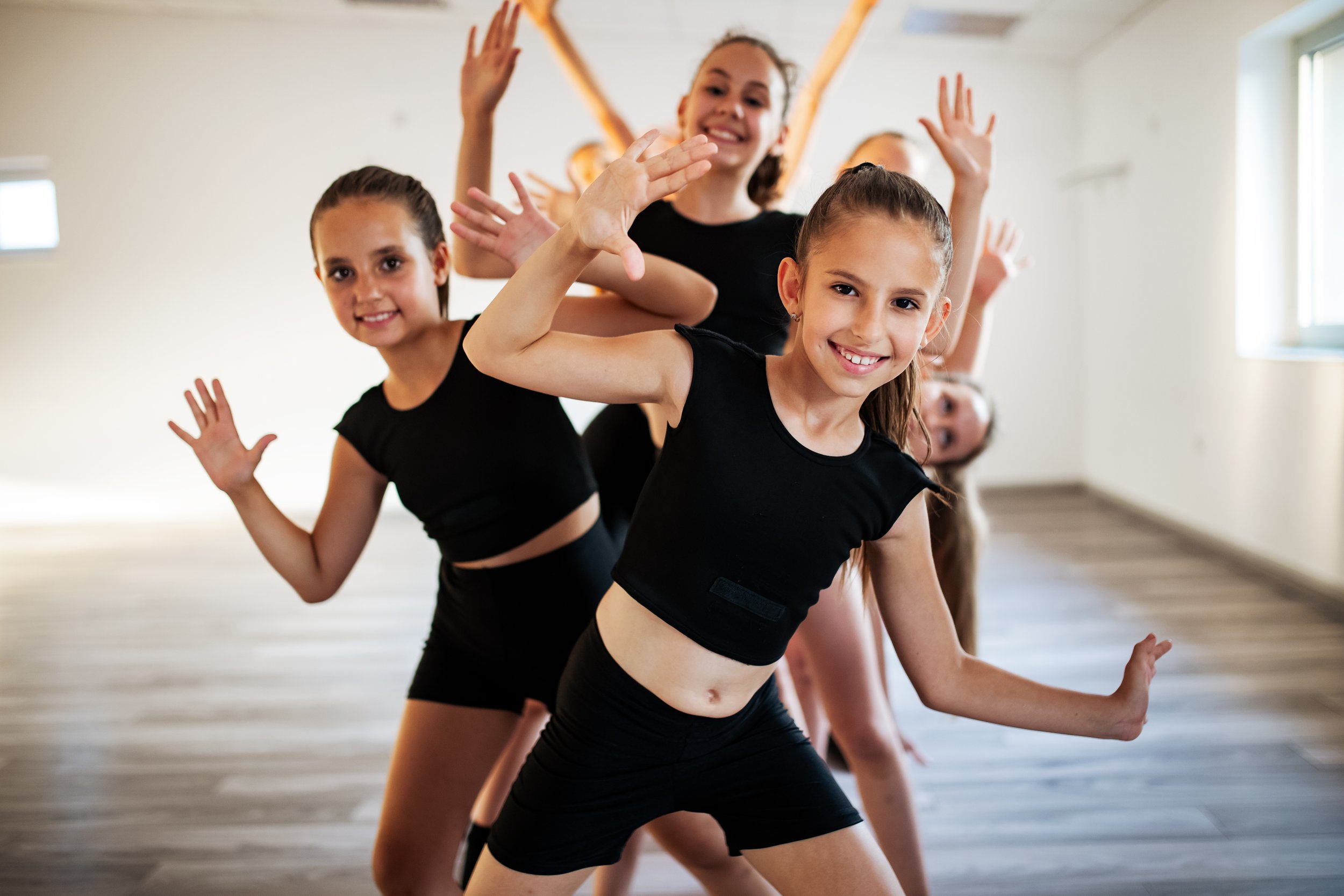 A group of young girls in black workout clothes practicing a dance or yoga pose in a bright studio, smiling and raising their hands.