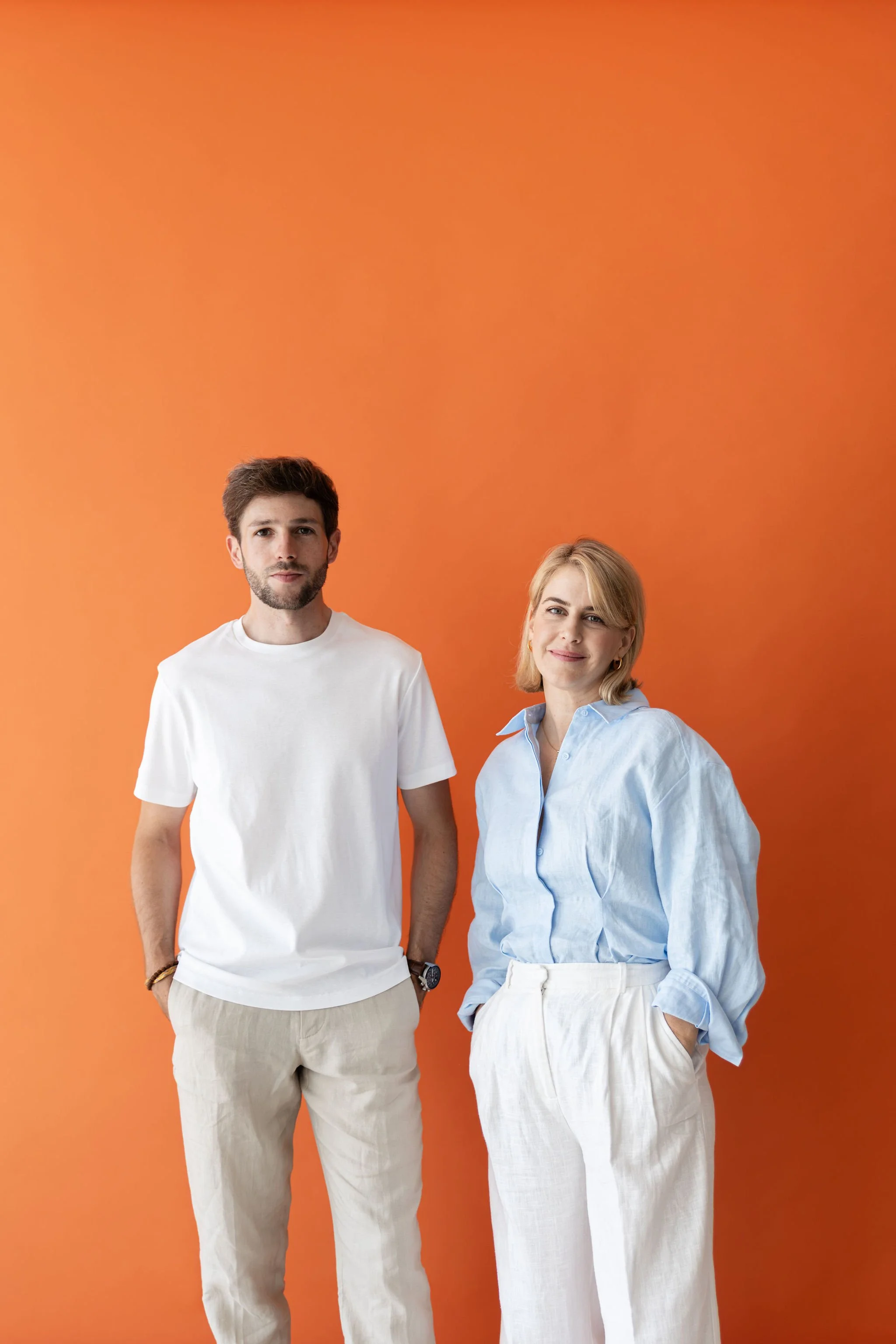 Un hombre y una mujer de pie frente a un fondo naranja. El hombre lleva una camiseta blanca y pantalones beige, y la mujer usa una blusa azul y pantalones blancos.