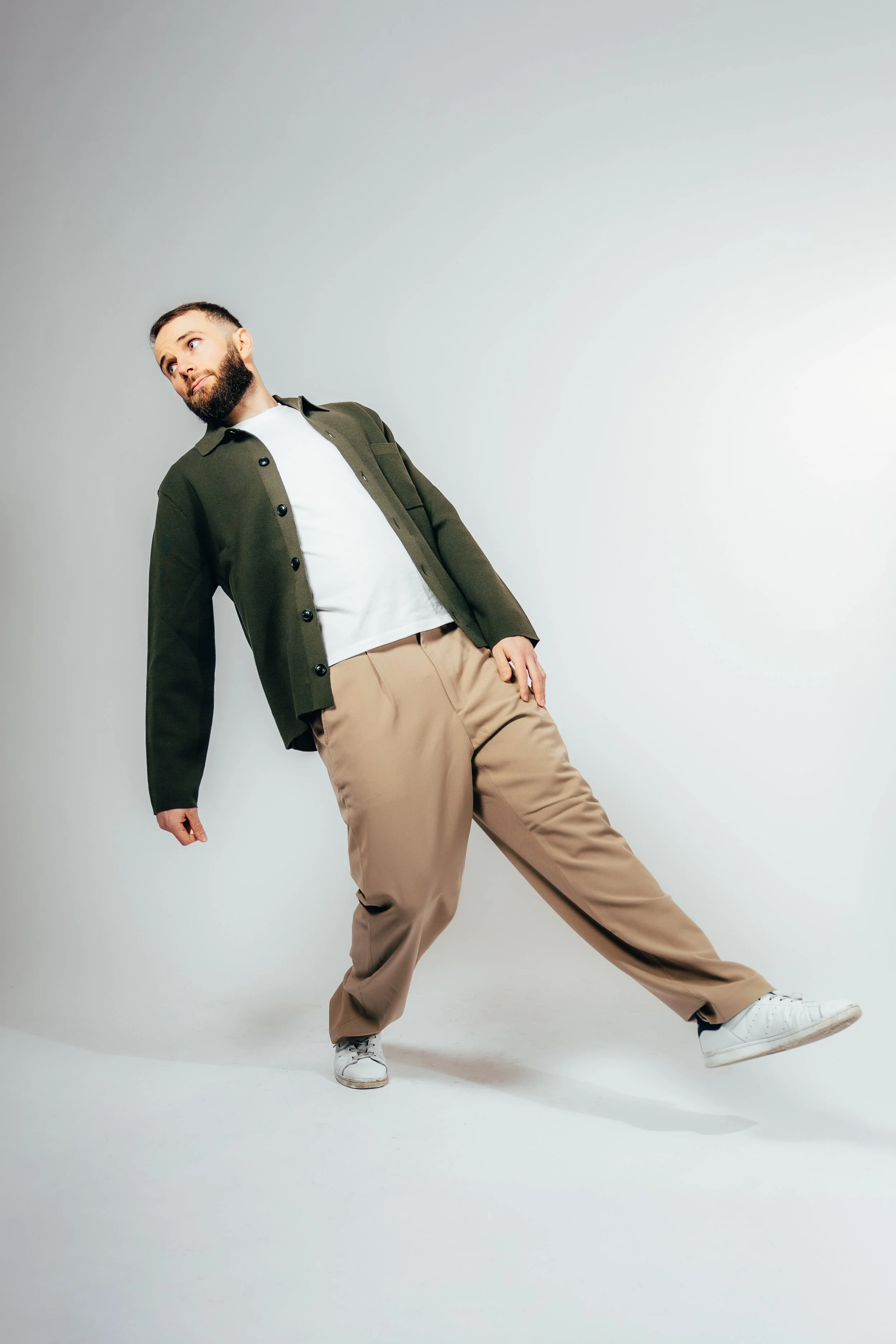 Jeune homme avec barbe, portant une veste kaki, un t-shirt blanc, un pantalon beige, et des chaussures blanches, en position décontractée contre un mur blanc.