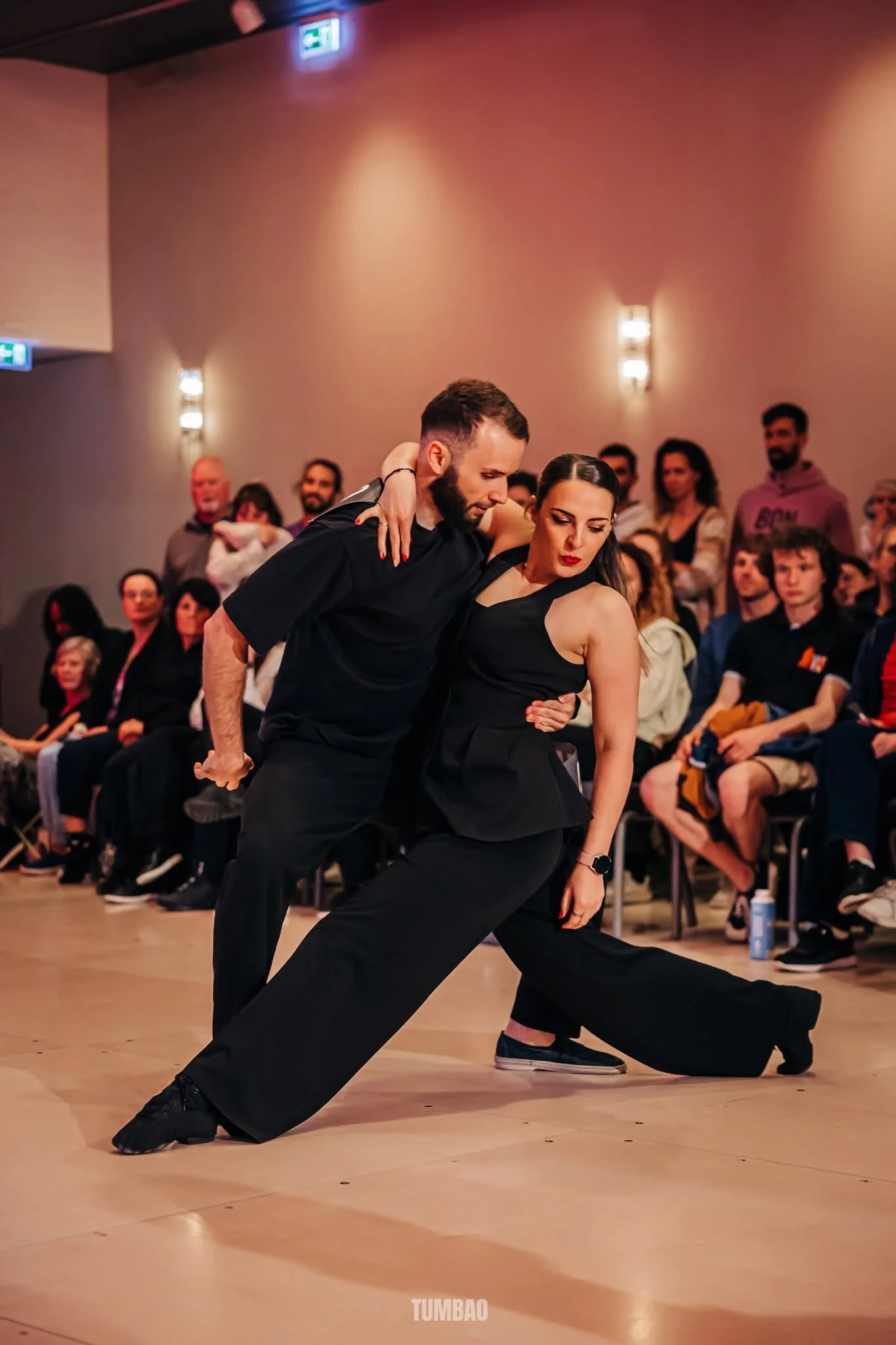 Deux danseurs en tenue noire exécutent une danse dans une salle avec un public spectateur.»}
