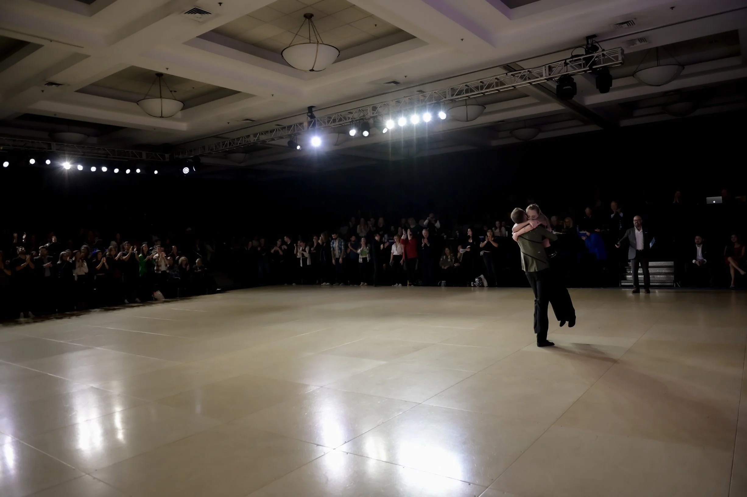Une scène d'une salle de bal ou d'un événement, avec un groupe de personnes qui semblent danser ou se préparer à danser, sous un éclairage puissant au plafond et entourée de spectateurs.