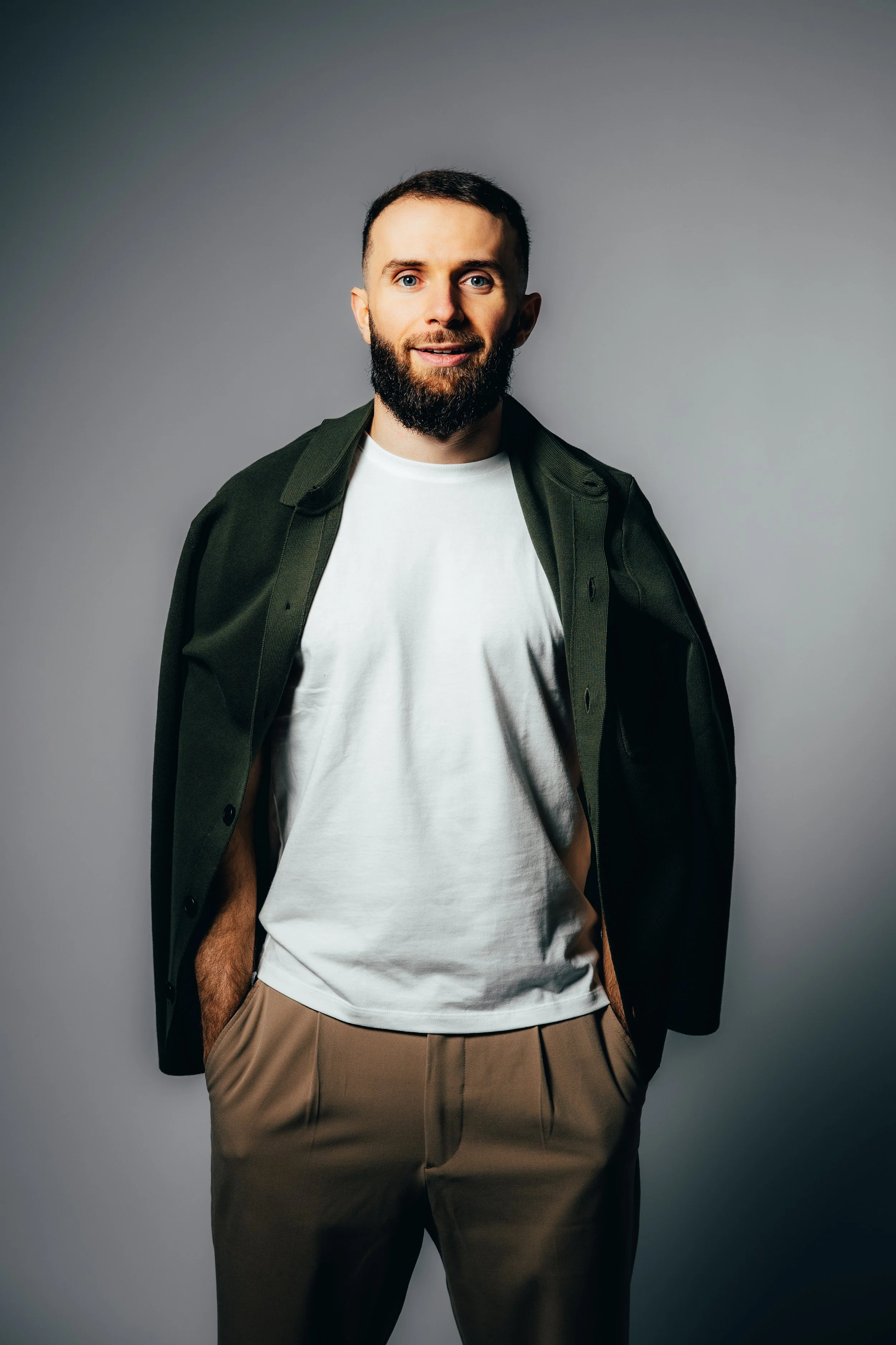 Homme debout portant un t-shirt blanc, un pantalon beige et une veste verte ouverte, souriant doucement, avec un fond gris