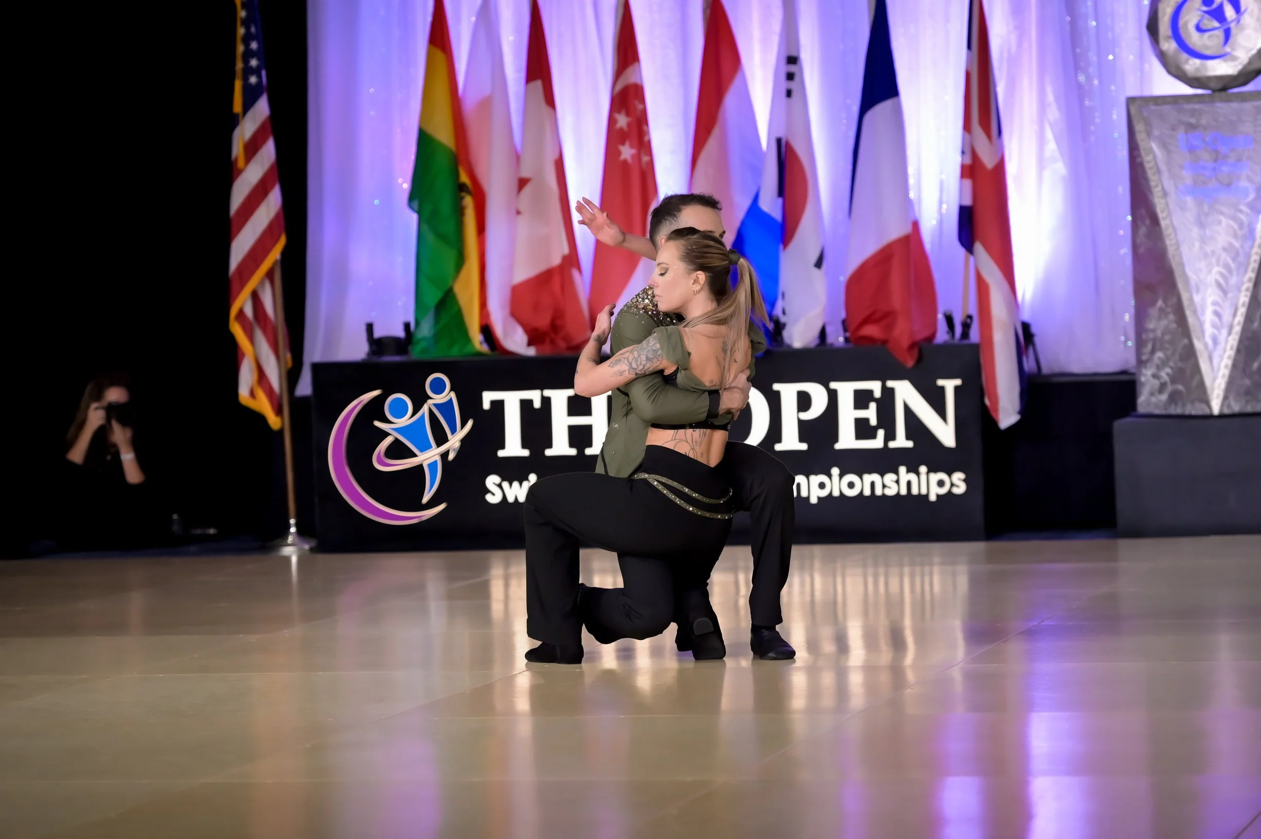 Deux danseurs effectuant une performance de danse contemporaine à l'intérieur d'une salle d'événements, devant un panneau avec le logo des Championnats du Monde de Danse (The Open). En arrière-plan, une rangée de drapeaux multicolores.