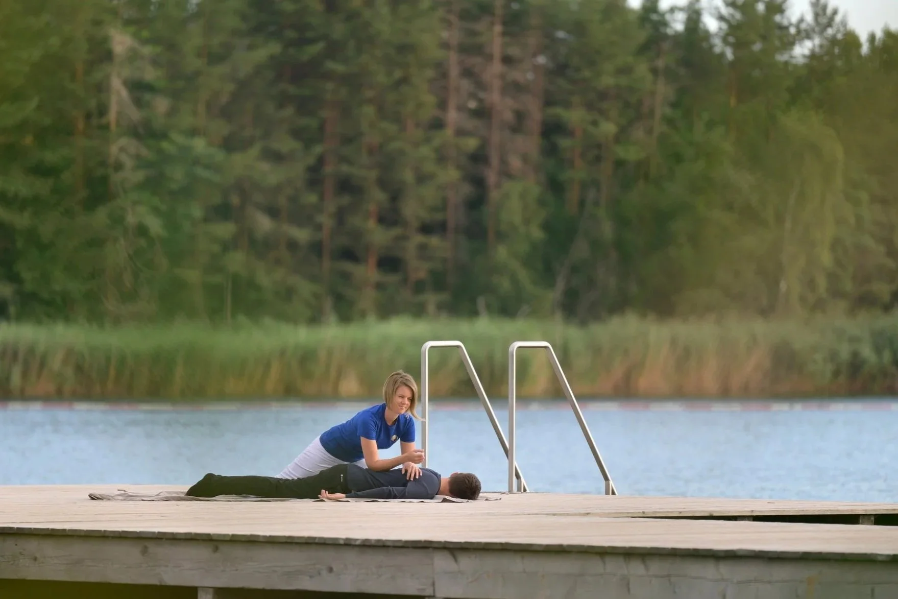 Eine Shiatsu Praktikerin führt eine Shiatsu Massage auf einer anderen Person durch, die auf dem Boden auf einer Matte liegt, am Ufer eines Sees mit einer Holzsteg und einer Leiter, im Hintergrund ein Wald.