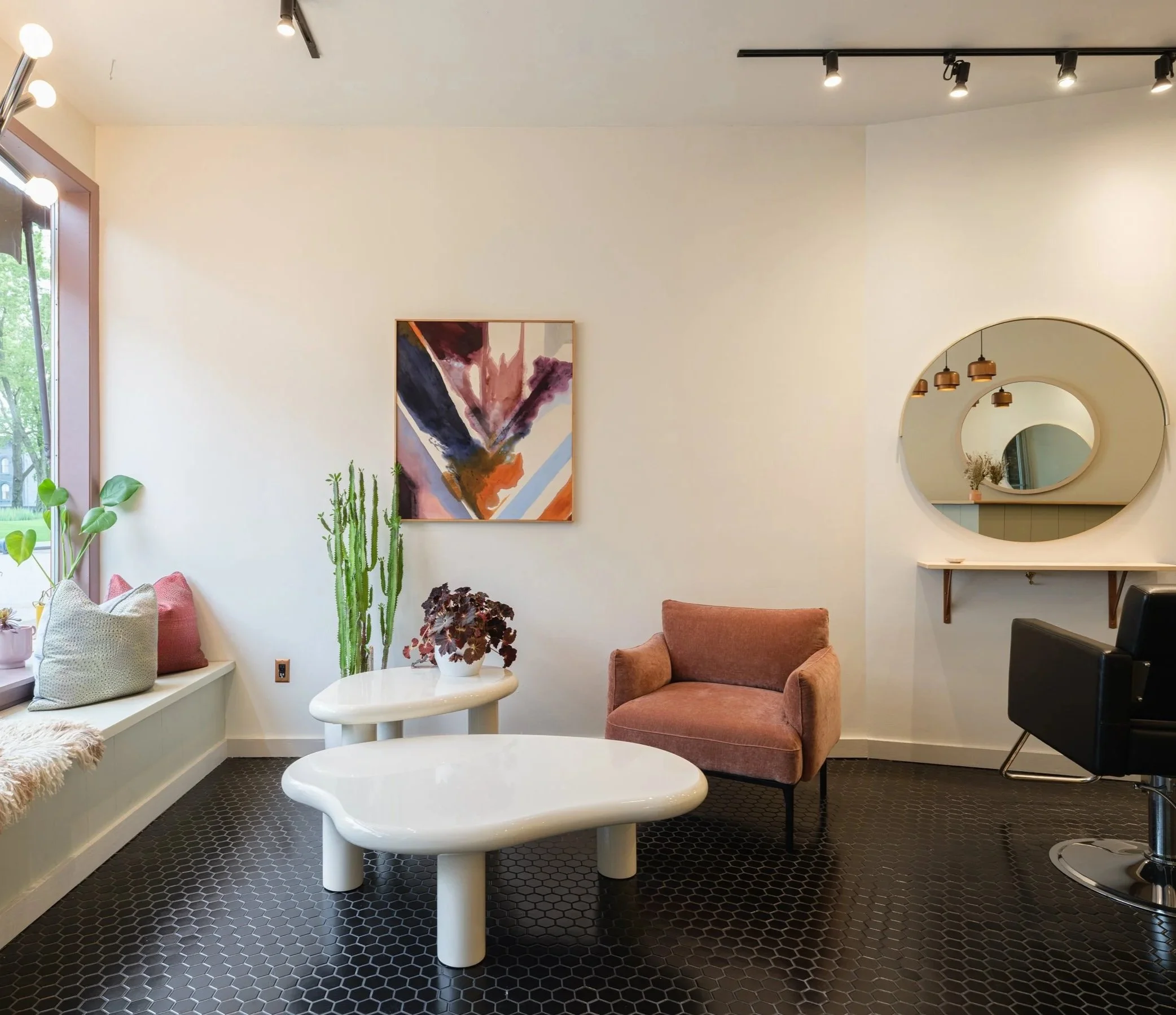 Interior of a modern room with a white wall, abstract artwork, a round mirror, a pink armchair, potted plants, and a window with blinds. Lighting includes ceiling spotlights.