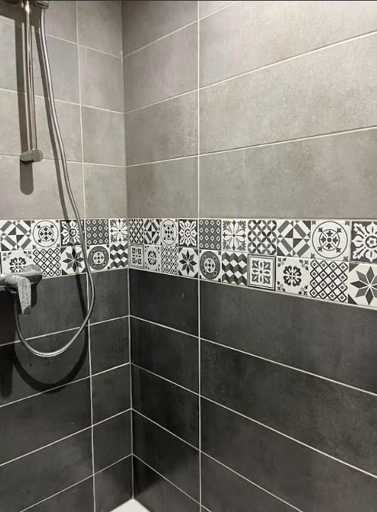 Carrelage de douche avec une rangée de carreaux décoratifs noirs, blancs et gris aux motifs variés, murs en grands carreaux gris clair et gris foncé, pommeau de douche en métal. Appartement situé à Montargis. Architecte d'intérieur Nadine Saguin,