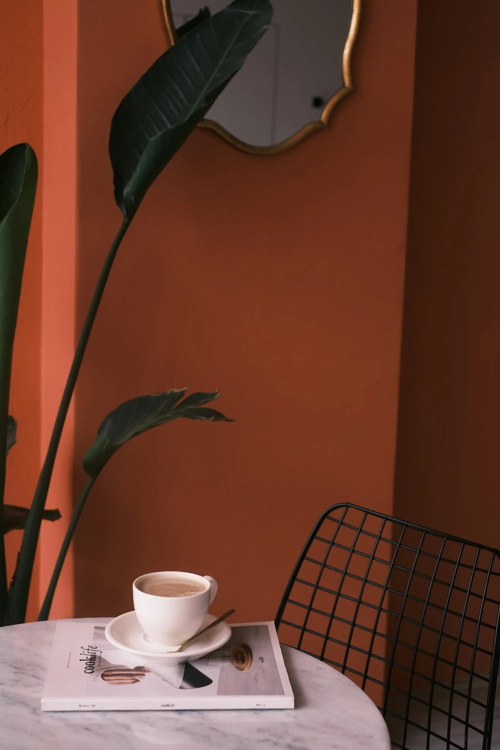 Une tasse de café sur une table en marbre avec un magazine ouvert dessous, une plante verte à gauche, un mur orange en arrière-plan, et une chaise en métal noir à droite.