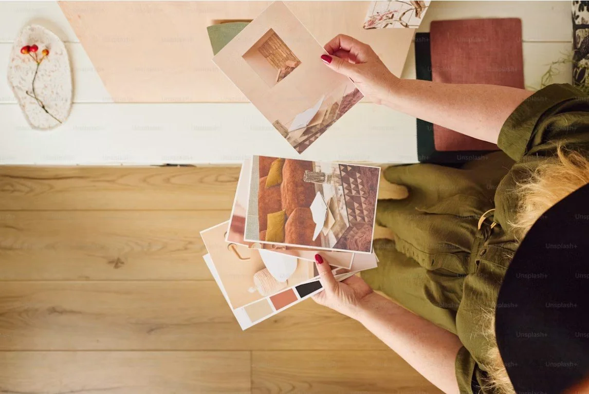 Une personne tenant plusieurs photos de décoration intérieure, avec un fond de table en bois et un mur blanc décoré d'une pièce en céramique