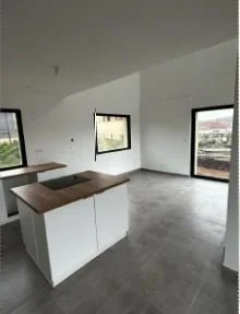 Intérieur d'une pièce avec deux fenêtres, murs blancs, sol en carrelage gris, cuisine avec comptoir en bois blanc. Projet d'aménagement conçu par Nadine Agencement & Design, architecte d'intérieure et décoratrice à Mions, Rhône.