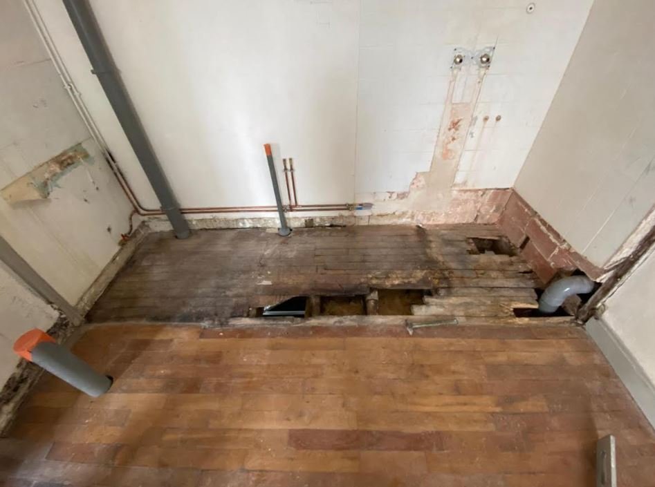 Une pièce en cours de rénovation, avec un sol partiellement enlevé révélant une structure en bois endommagée et des tuyaux apparents.