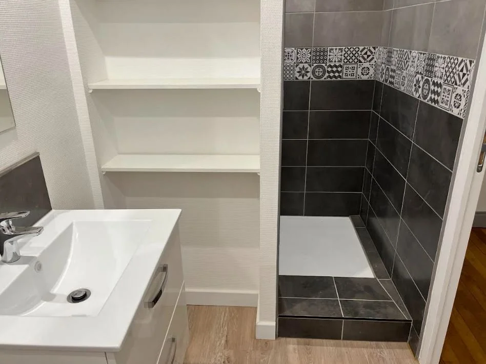 Salle de bain avec lavabo blanc, étagère ouverte et zone de douche avec carrelage gris et décoratif. projet conçu par Nadine Agencement & Design, architecte d'intérieure et décoratrice à Mions, Rhône
