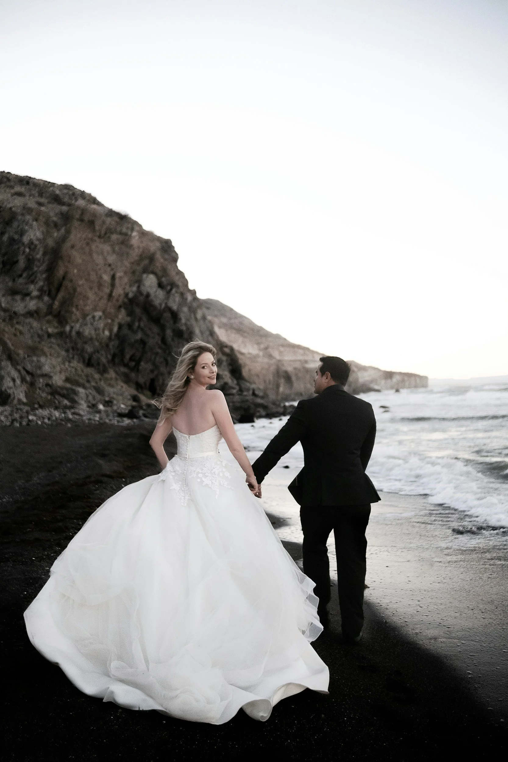 A couple in wedding attire standing on a black sand beach near rocky cliffs, with ocean waves in the background, during sunset.