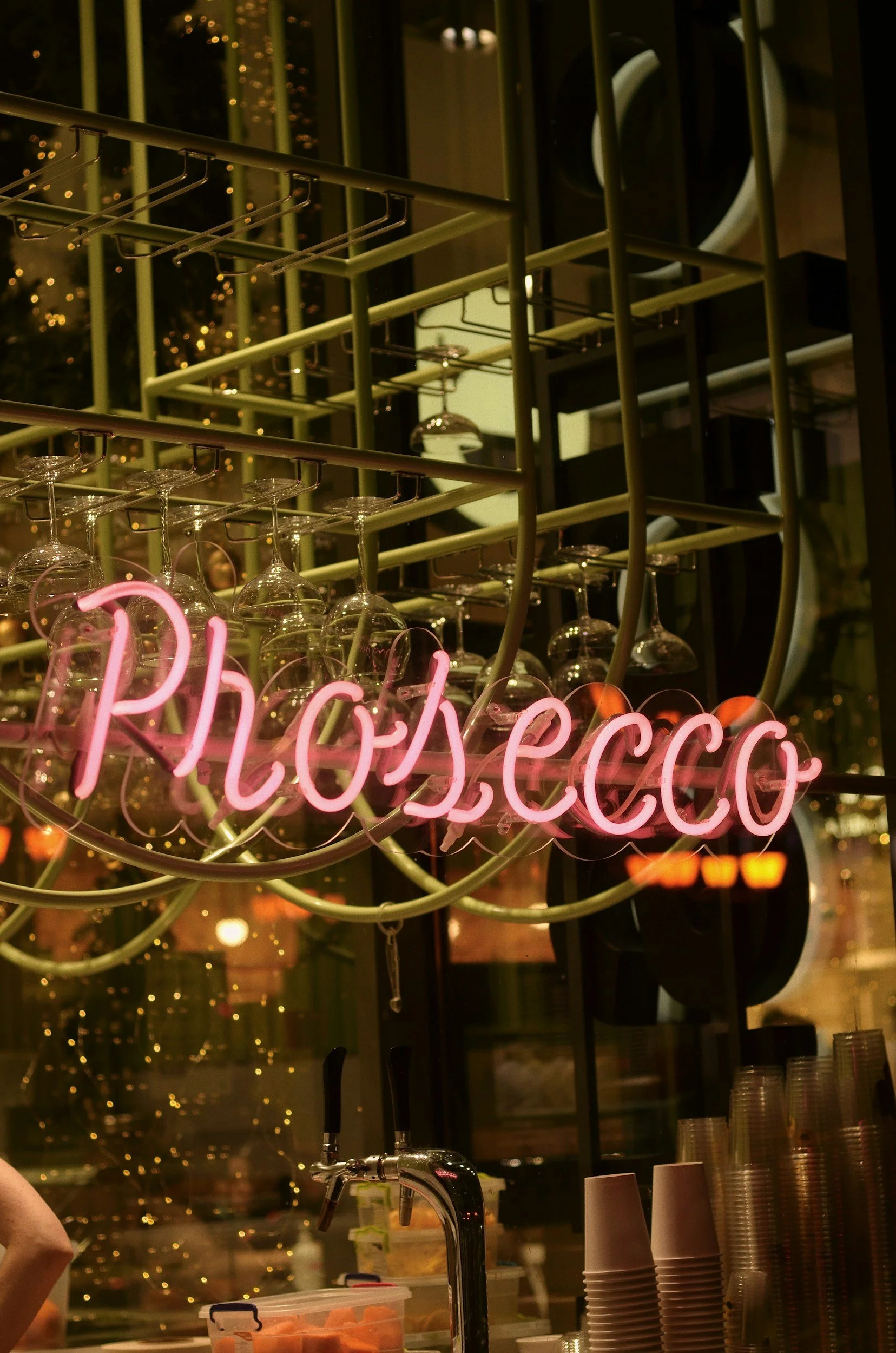 Close-up of a neon pink sign that spells out 'Prosecco' hanging between wine glasses and a bar counter, with a blurred background of a bar.