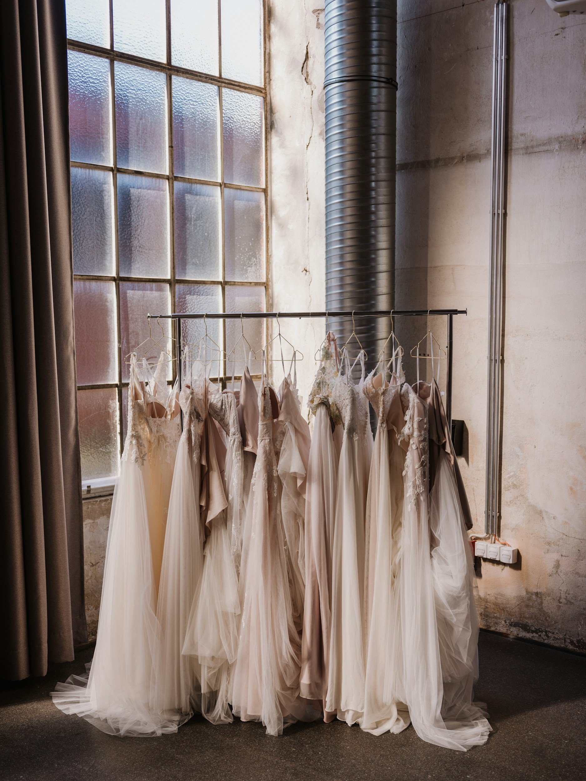 Multiple wedding dresses hanging on a clothing rack in front of a large, textured glass window with black curtains on the side and a metal air duct in the background.