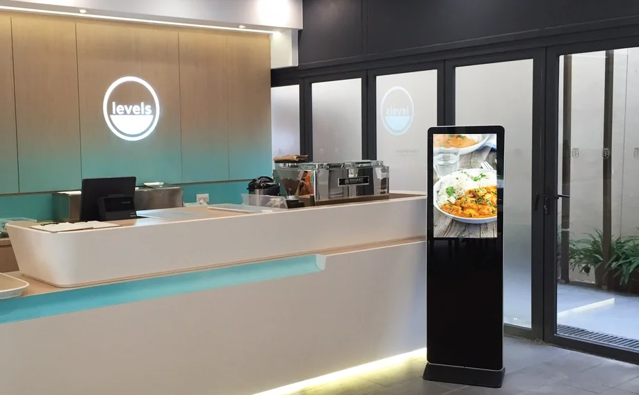 Modern cafe or restaurant counter with a digital order kiosk displaying a plate of food, behind the counter are beverage machines and a cash register, with the logo 'levels' illuminated on the wall.