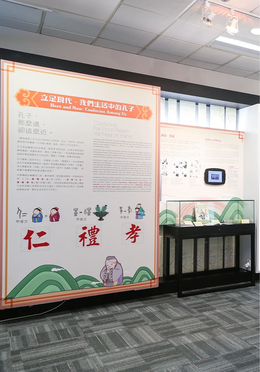 An exhibition display panel titled 'Here and Now: Confucius Amongst Us' with text in Chinese and English, illustrations of Confucius and traditional Chinese symbols, and a glass display case with brochures or pamphlets in front of a white wall with d