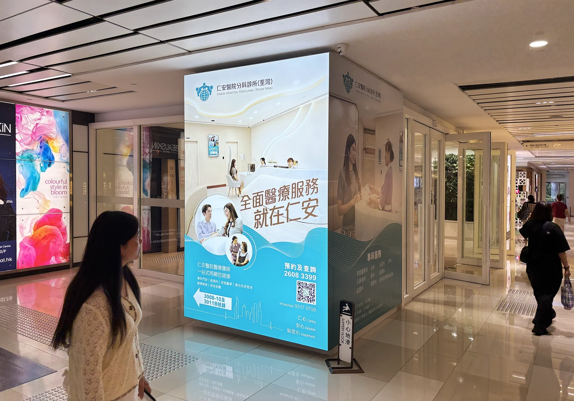 Digital advertisement for Union Hospital Polyclinic in Tsuen Wan, featuring images of medical staff and contact information. The scene is inside an indoor shopping mall with people walking by.